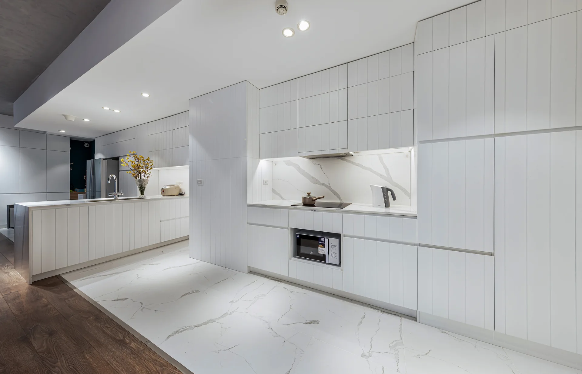 A modern minimalist white kitchen with vertical-slatted cabinets, veined marble countertops, and under-cabinet lighting.