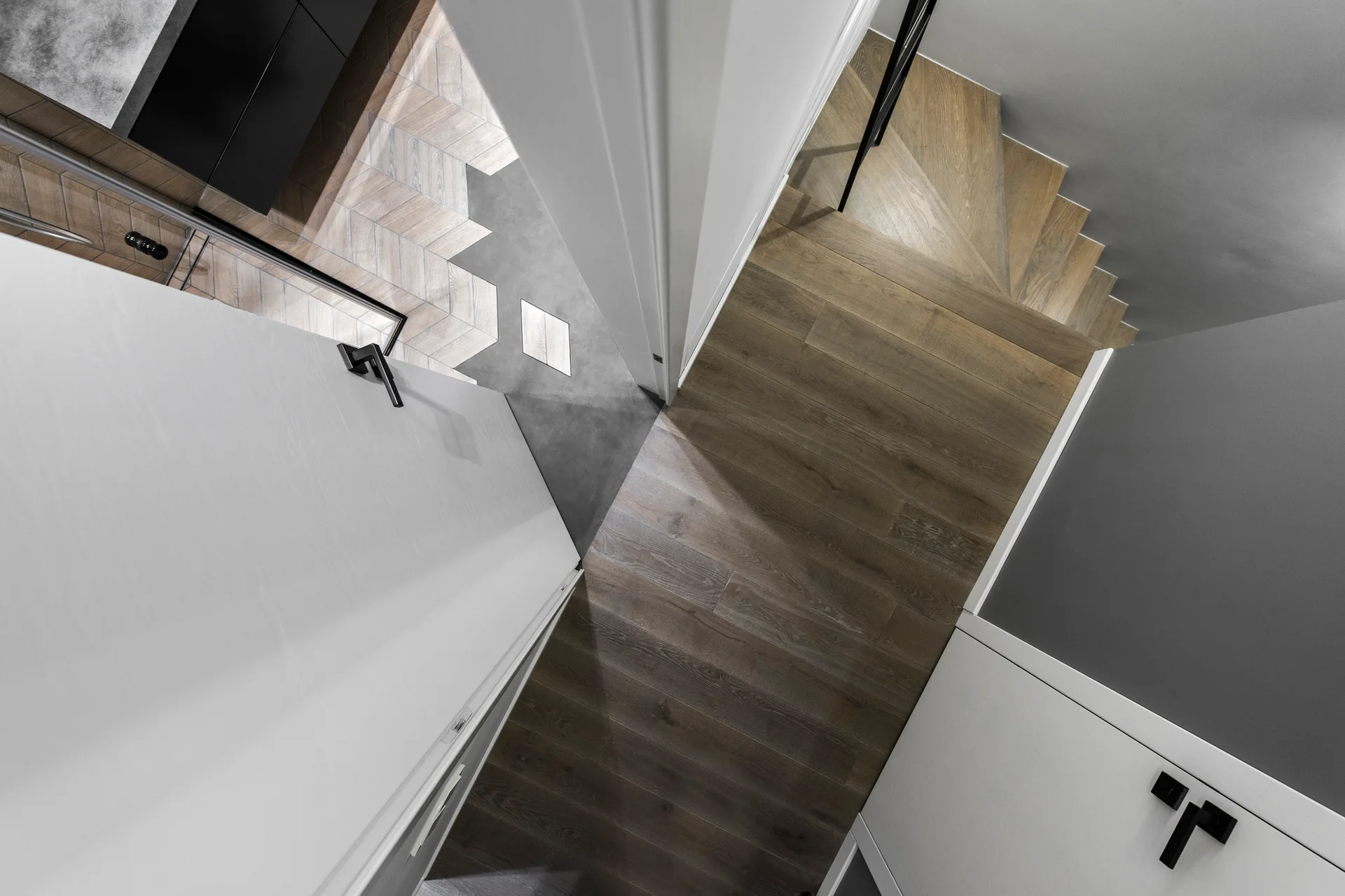 A high-angle view of a modern hallway showing a wooden staircase, white doors, and a geometric transition from wood planks to grey tiles.