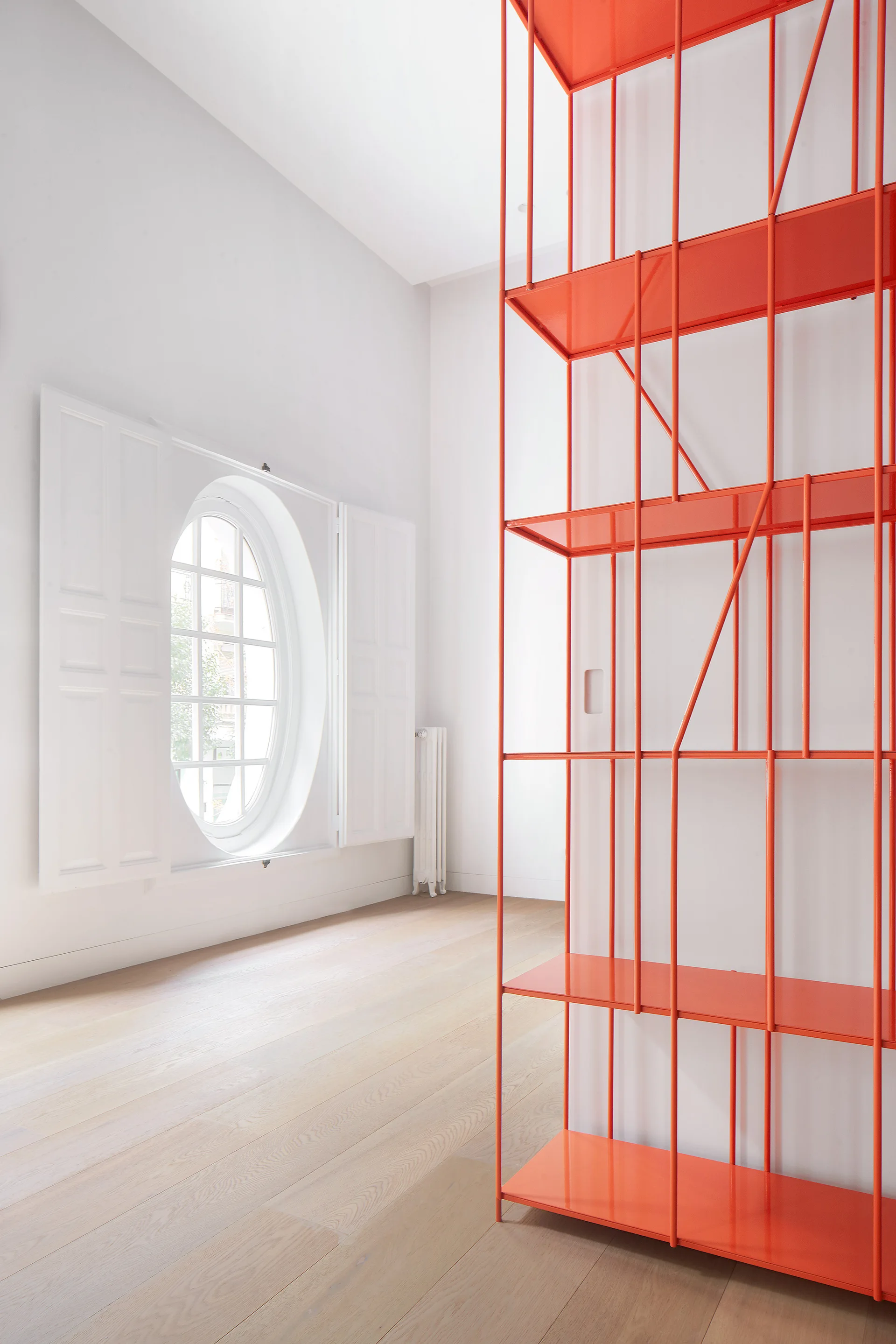 A bright minimalist room with light wood floors features a tall, glossy orange metal shelving unit and a white oval window with shutters.