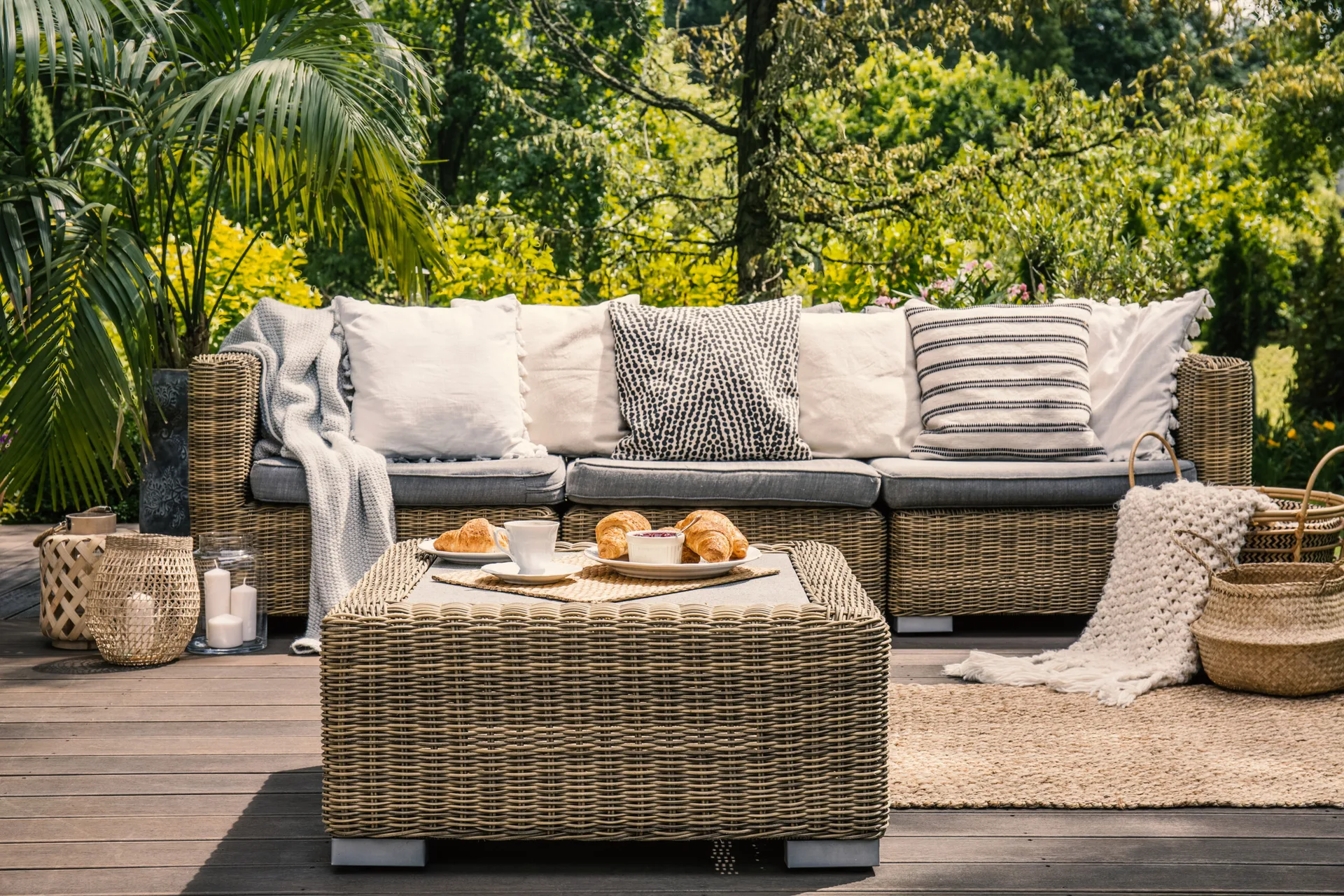 A wicker outdoor sofa and matching coffee table set on a wooden deck with throw pillows, knit blankets, and lanterns against a lush garden backdrop.