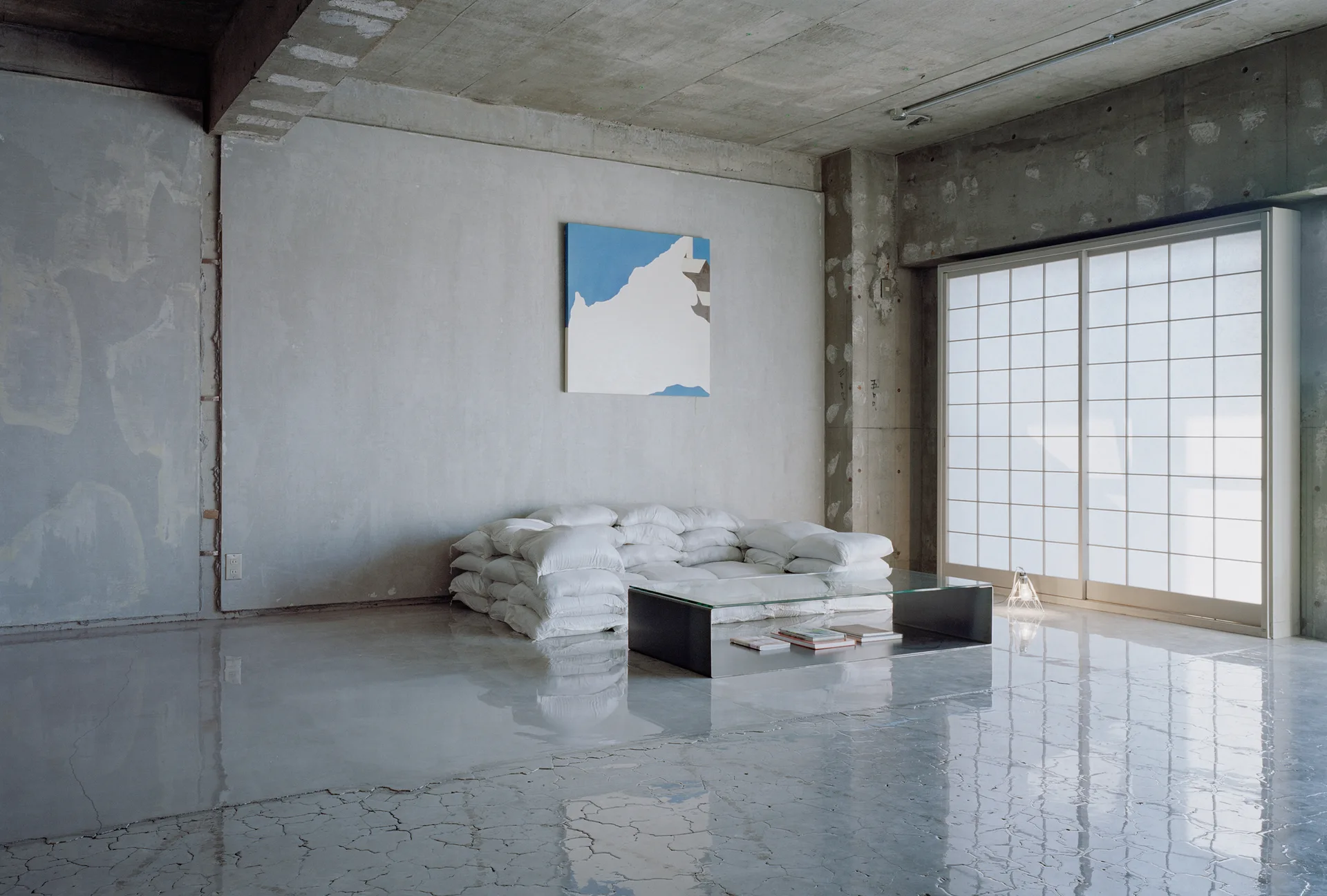 An industrial living room with raw concrete walls, a polished floor, a stacked white cushion sofa, and a glass coffee table.