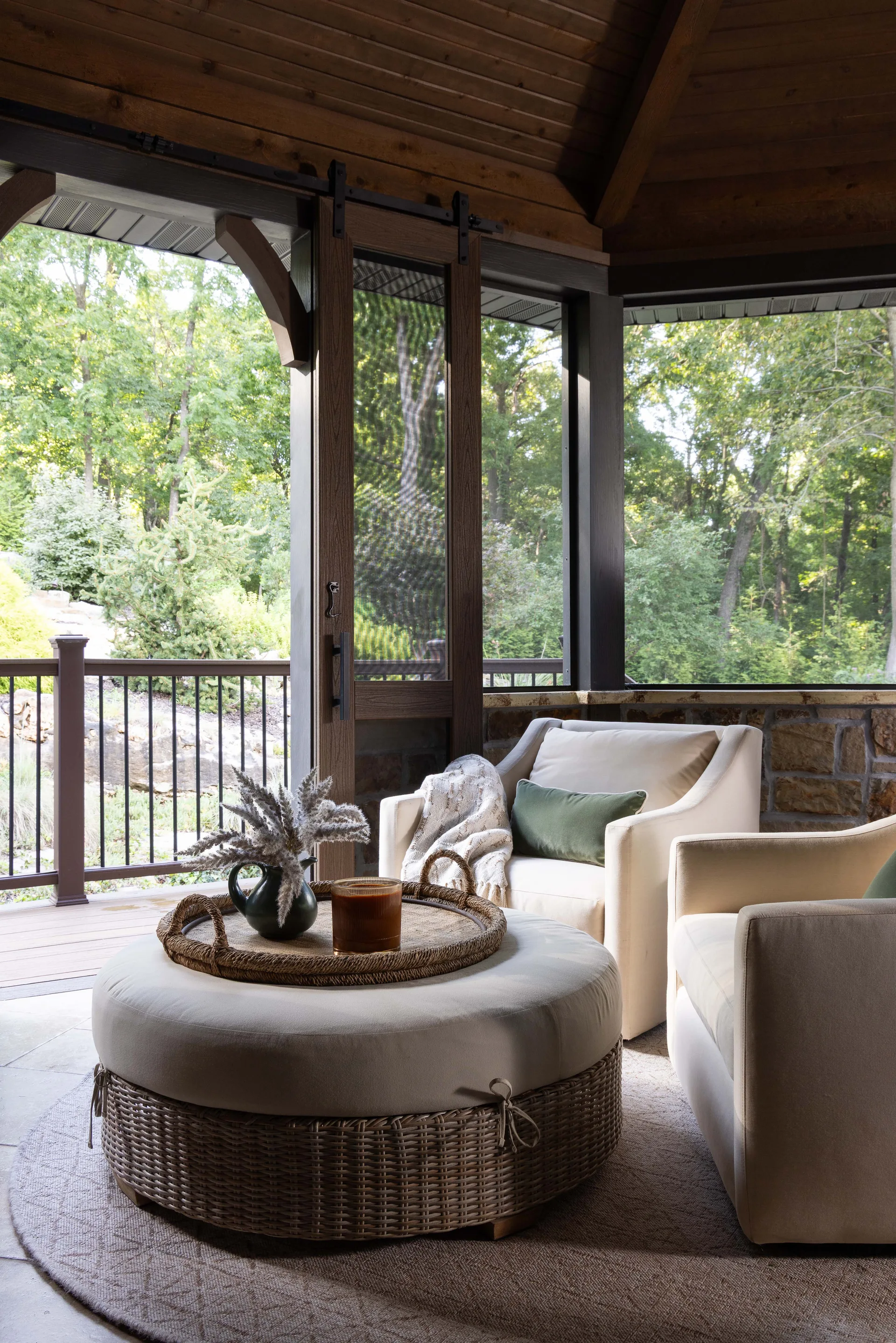 Two beige upholstered armchairs and a round wicker-base ottoman sit on a circular rug inside a screened porch with a wood ceiling.