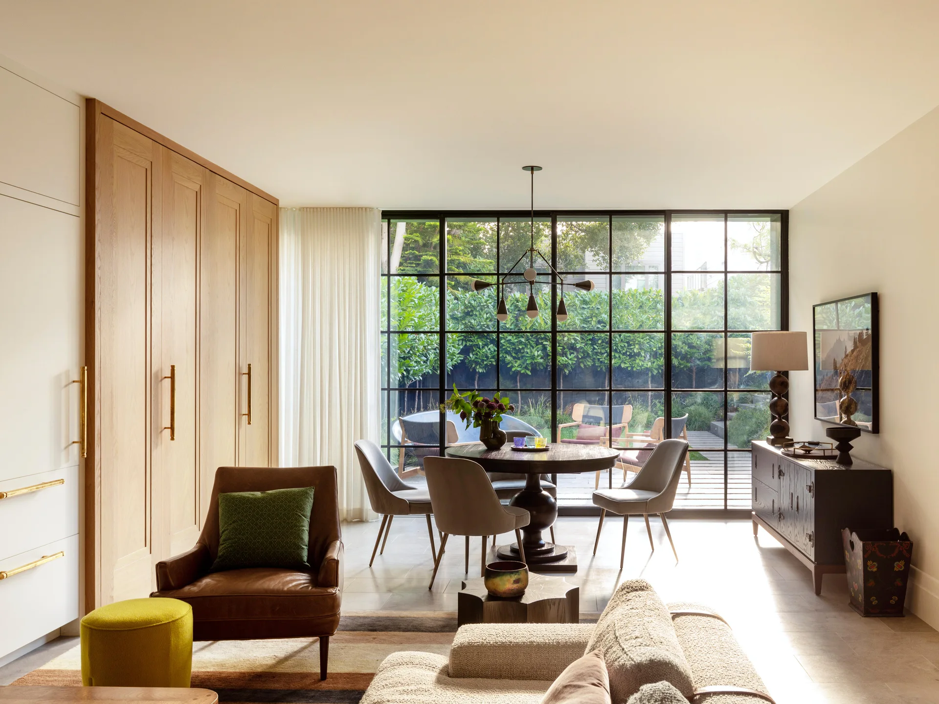 A contemporary living and dining room with a light oak cabinetry wall, a round dining table, and a large black metal grid window.