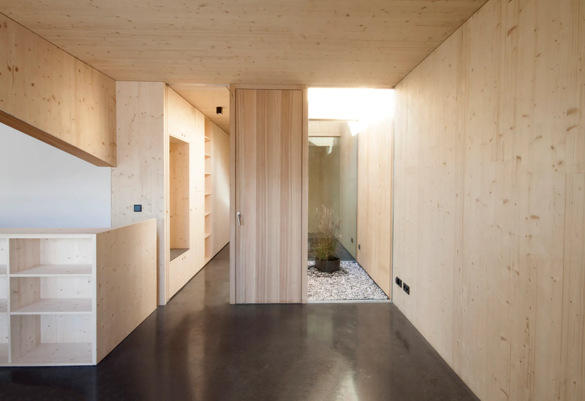A minimalist hallway features light wood paneled walls and ceiling, a dark concrete floor, and an internal glass-enclosed gravel courtyard.