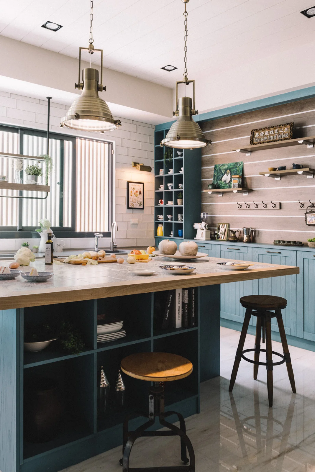 A kitchen features a teal island with integrated open shelving, a wood countertop, matching cabinetry, and industrial brass pendant lights.
