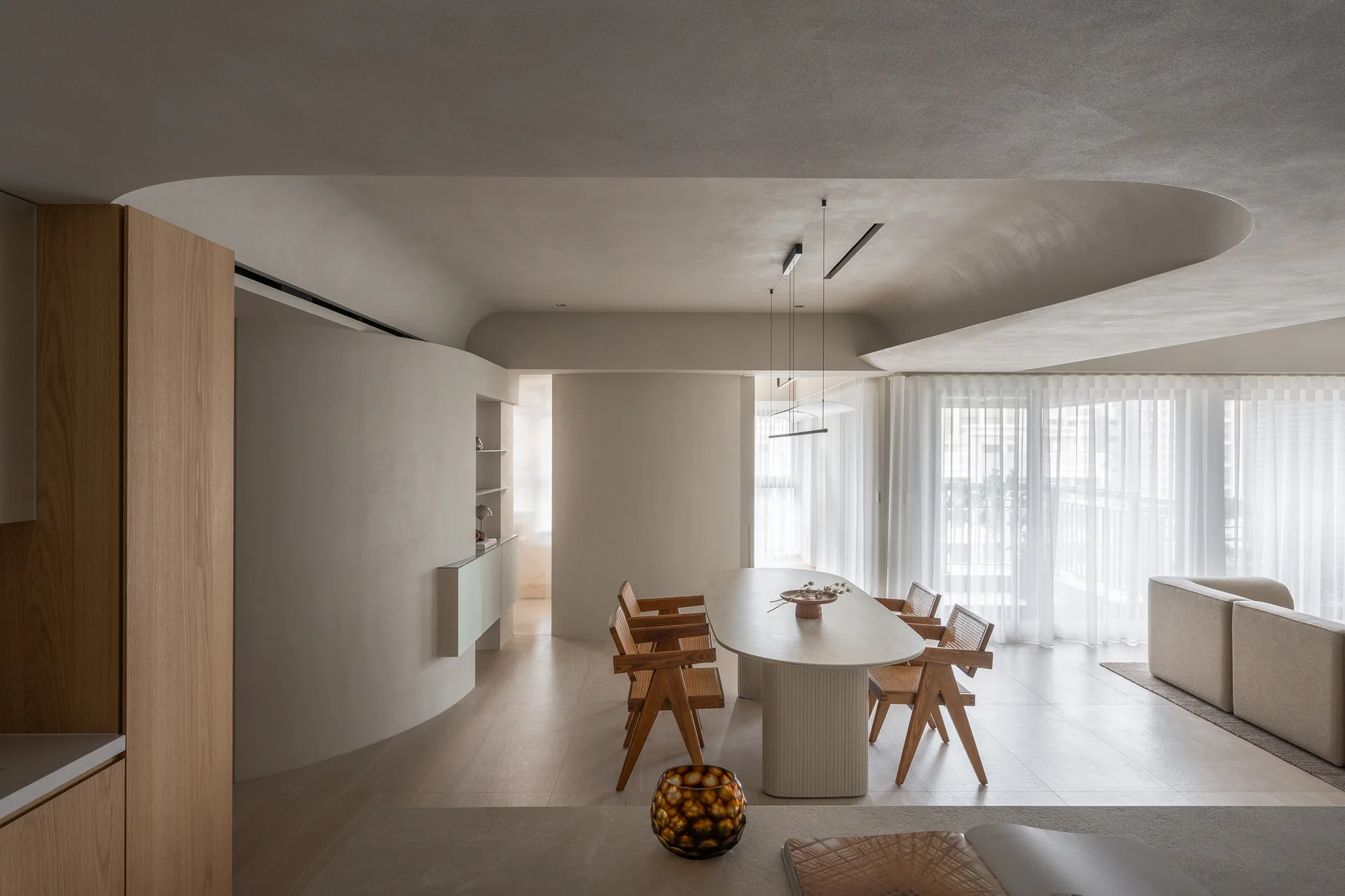A minimalist dining room features an oval table with cane-back wooden chairs set against a curved plaster wall and sheer curtains.