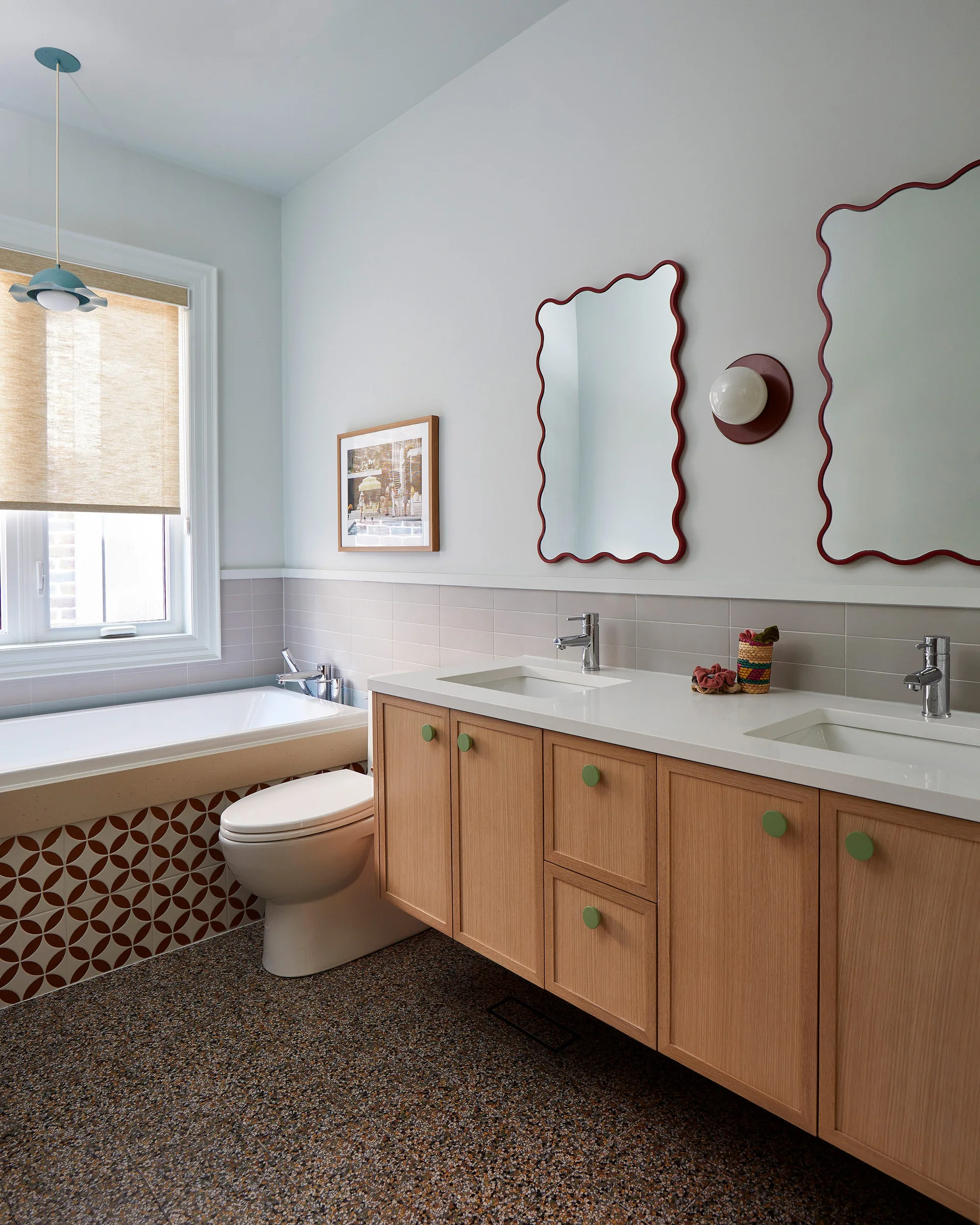 A modern bathroom features a floating light wood vanity, two red scalloped mirrors, a built-in tub with geometric tile, and a speckled floor.