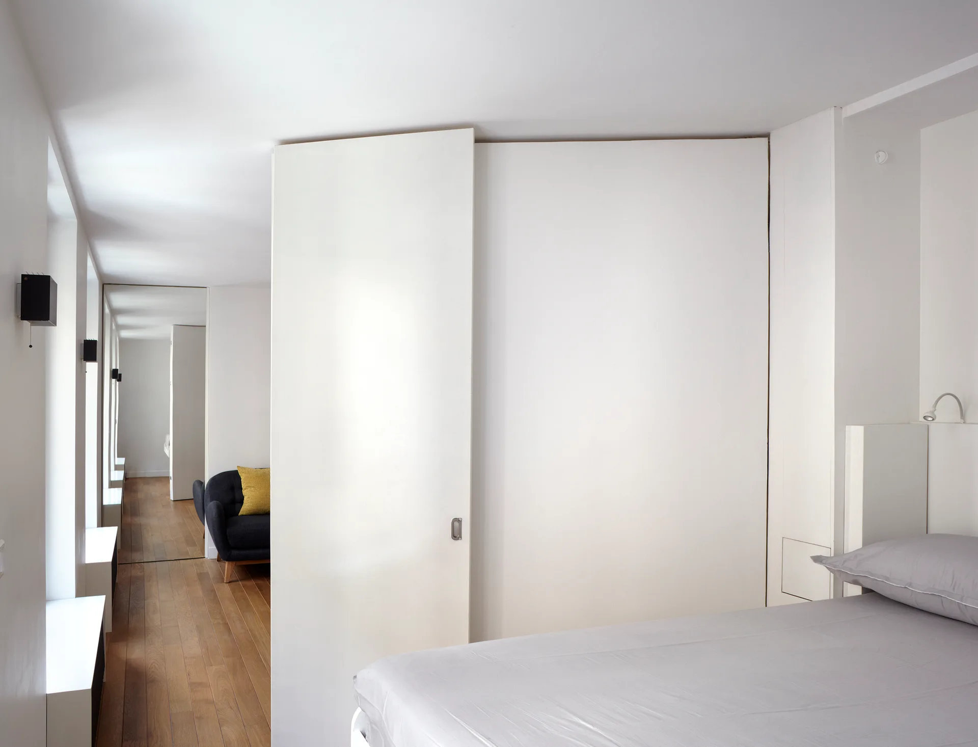 A minimalist bedroom featuring grey bedding, a large white sliding partition door, and a view into a hallway with hardwood flooring.