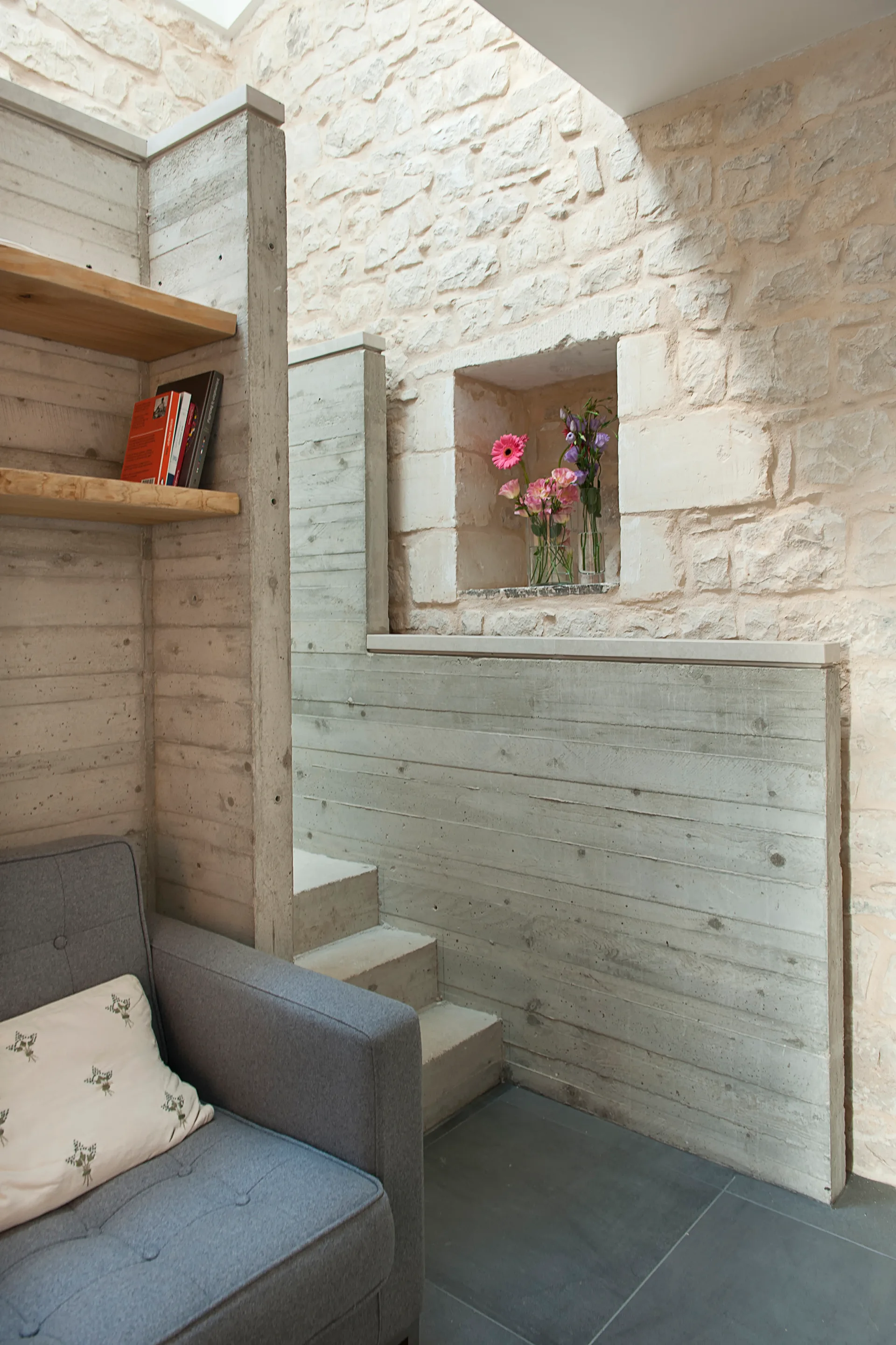 A grey armchair sits next to a board-marked concrete divider wall and a rustic stone wall featuring a recessed niche with flowers.