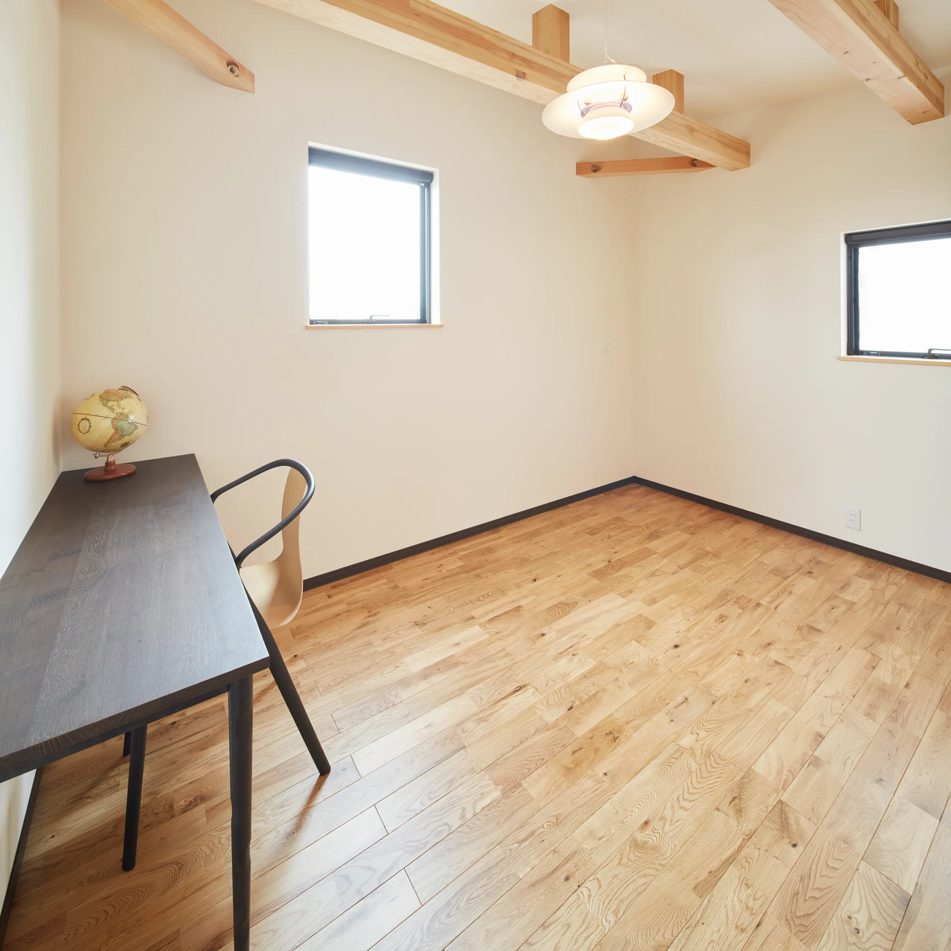A minimalist home office features exposed light wood ceiling beams, hardwood flooring, cream walls, and a dark brown desk with a curved wooden chair.
