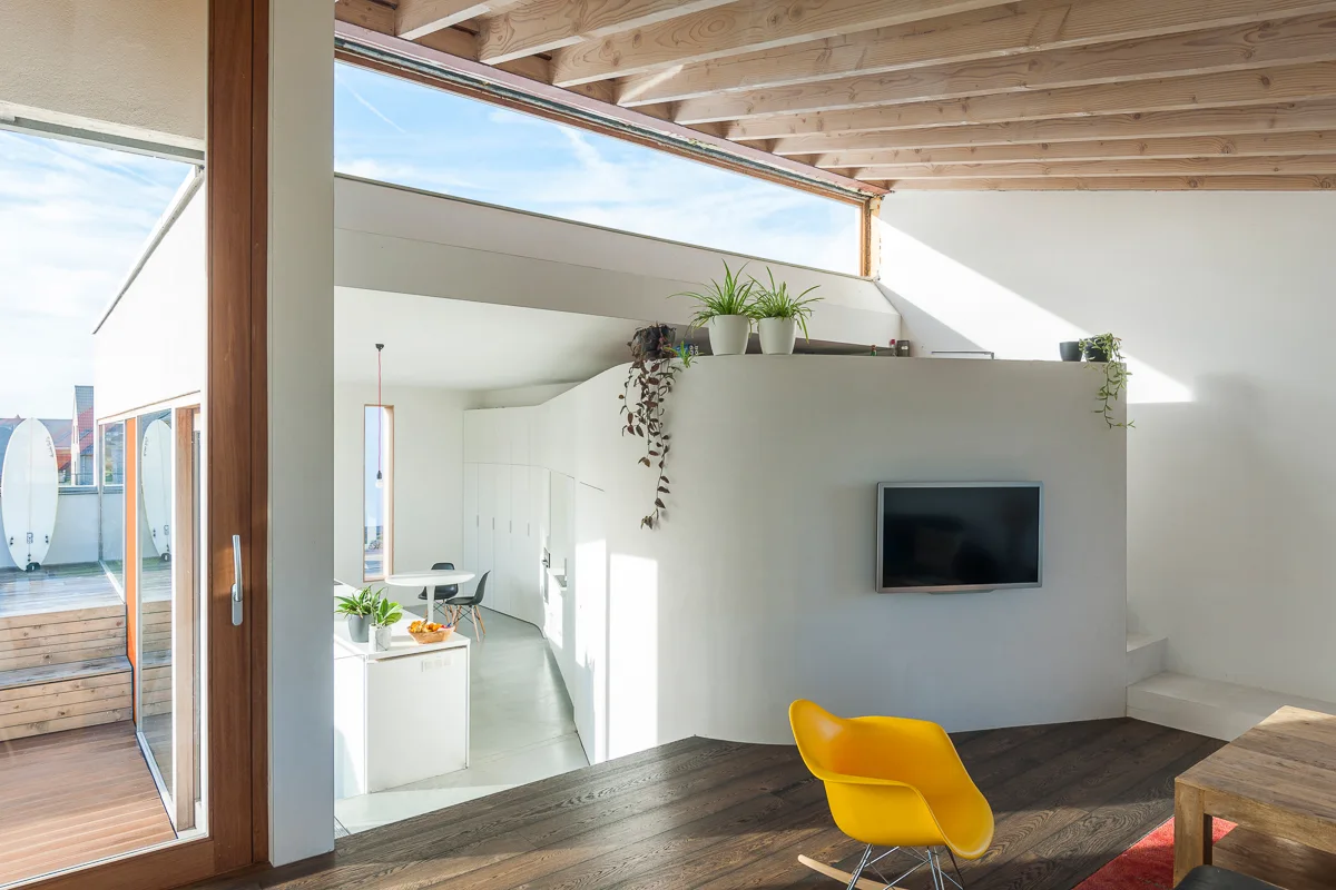 A modern open-plan living room featuring a curved white partition wall, exposed wood ceiling rafters, and a bright yellow rocking chair.