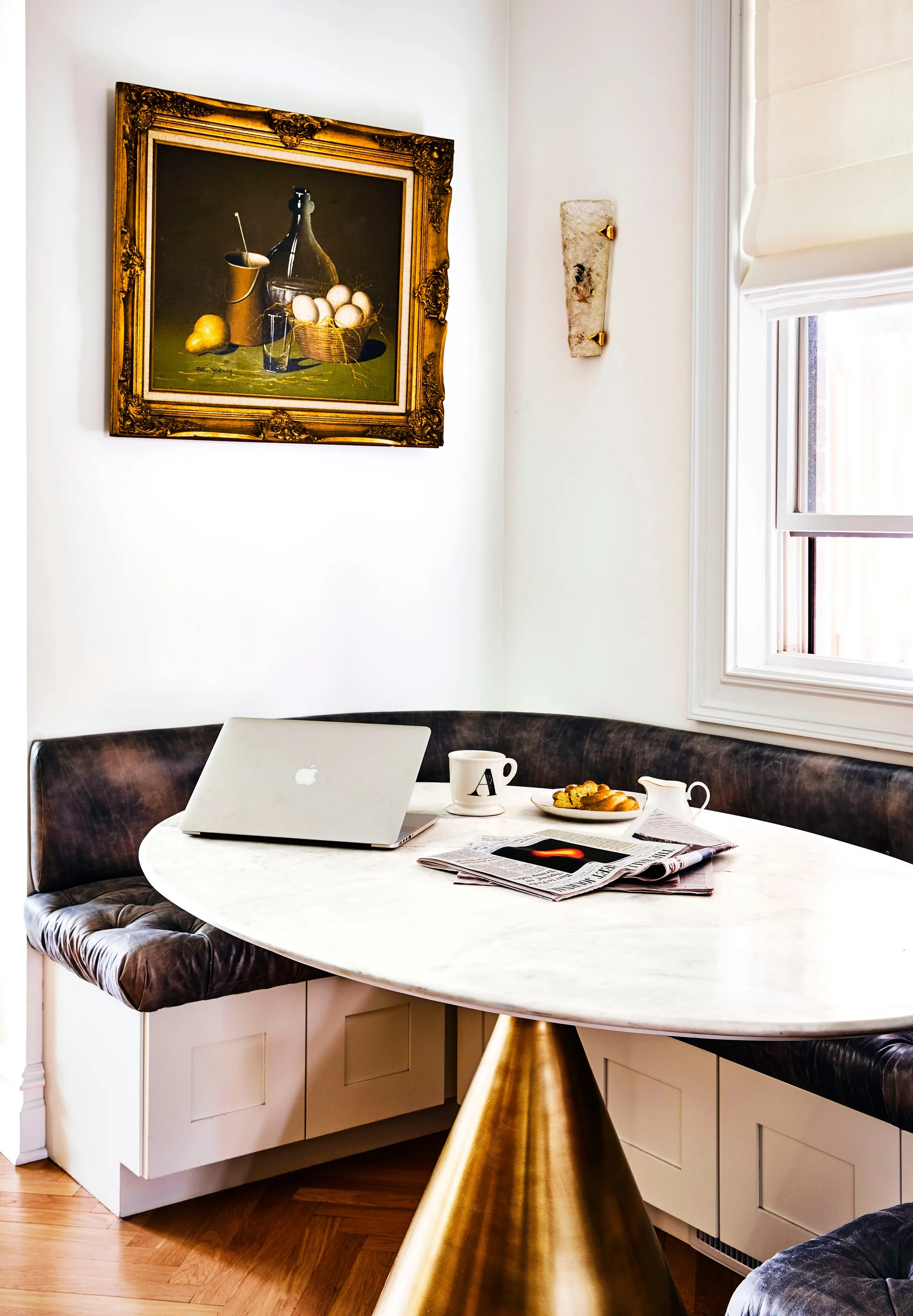 A breakfast nook features a curved tufted leather banquette and an oval marble table with a conical brass pedestal base beneath a framed oil painting.