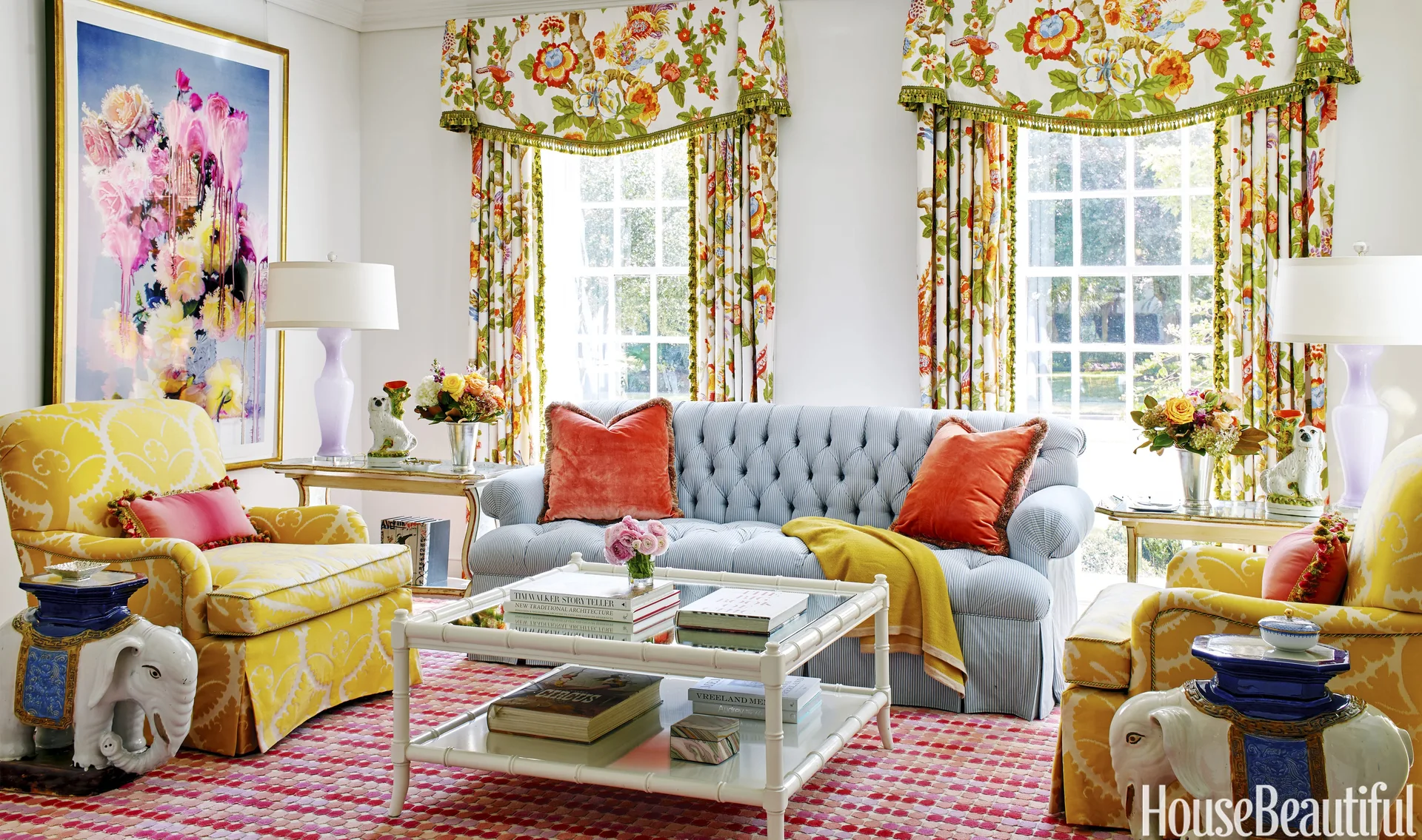 A maximalist living room features a tufted blue striped sofa, yellow patterned armchairs, floral print curtains, and a white bamboo coffee table.