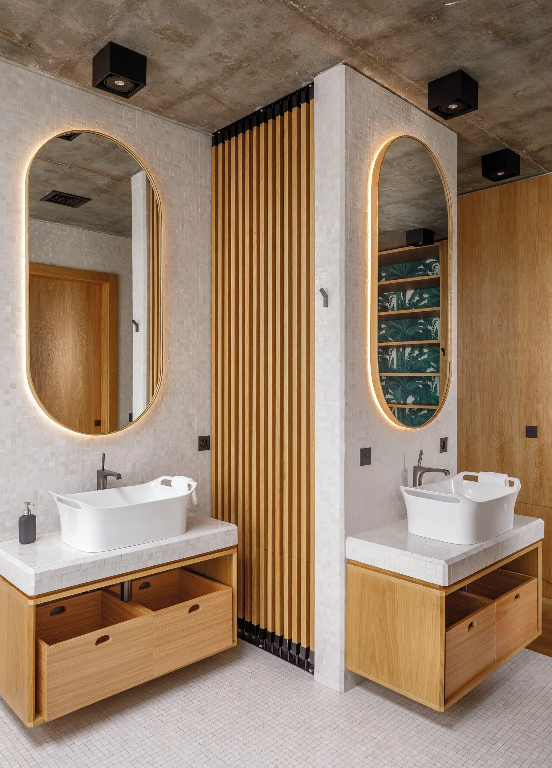 A modern bathroom featuring two separate wood vanities with white vessel sinks, oval backlit mirrors, and a vertical wood slat partition against mosaic tile.