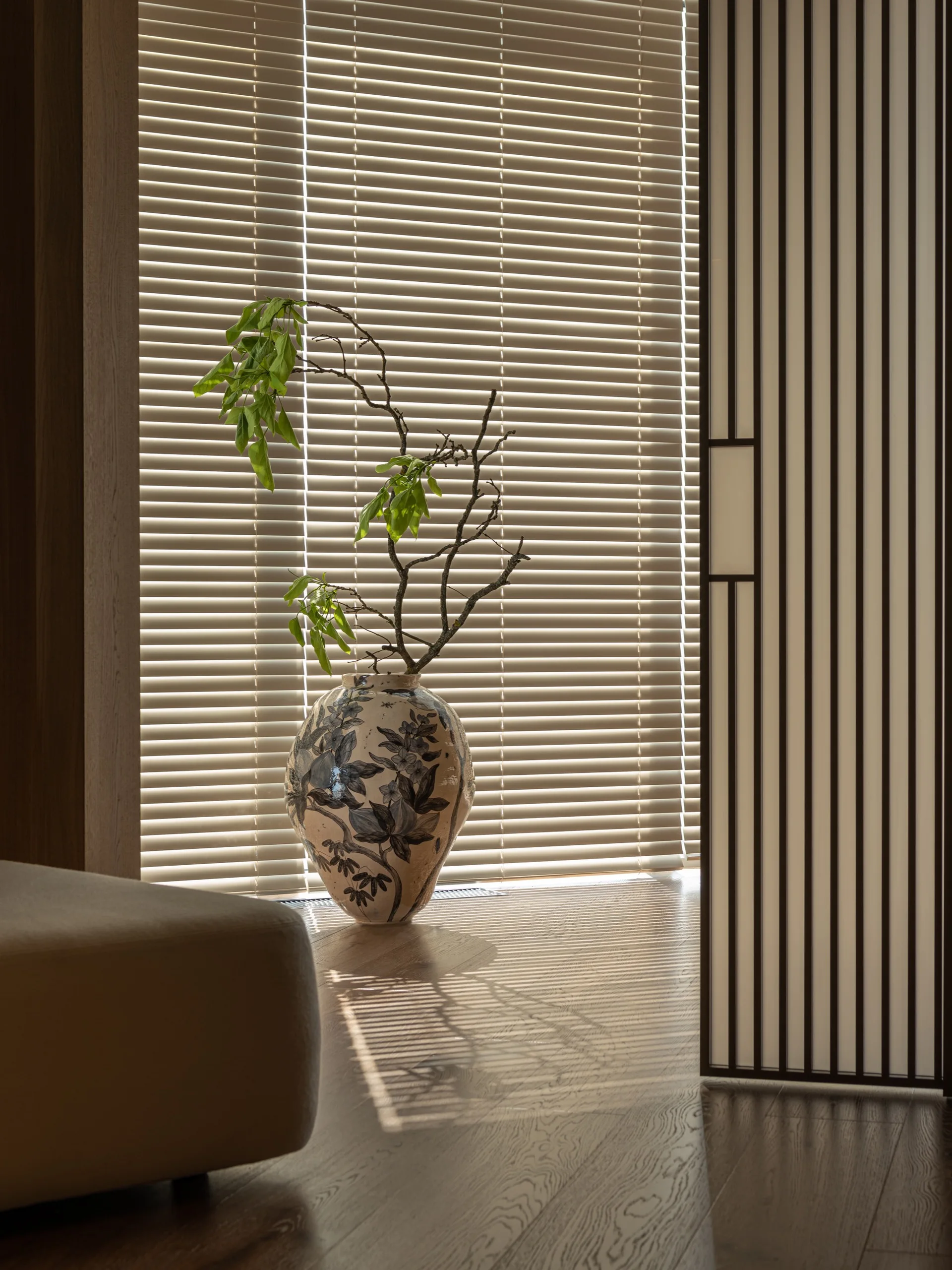 A hand-painted ceramic vase holding leafy branches sits on a hardwood floor next to horizontal window blinds and a vertical slatted screen.
