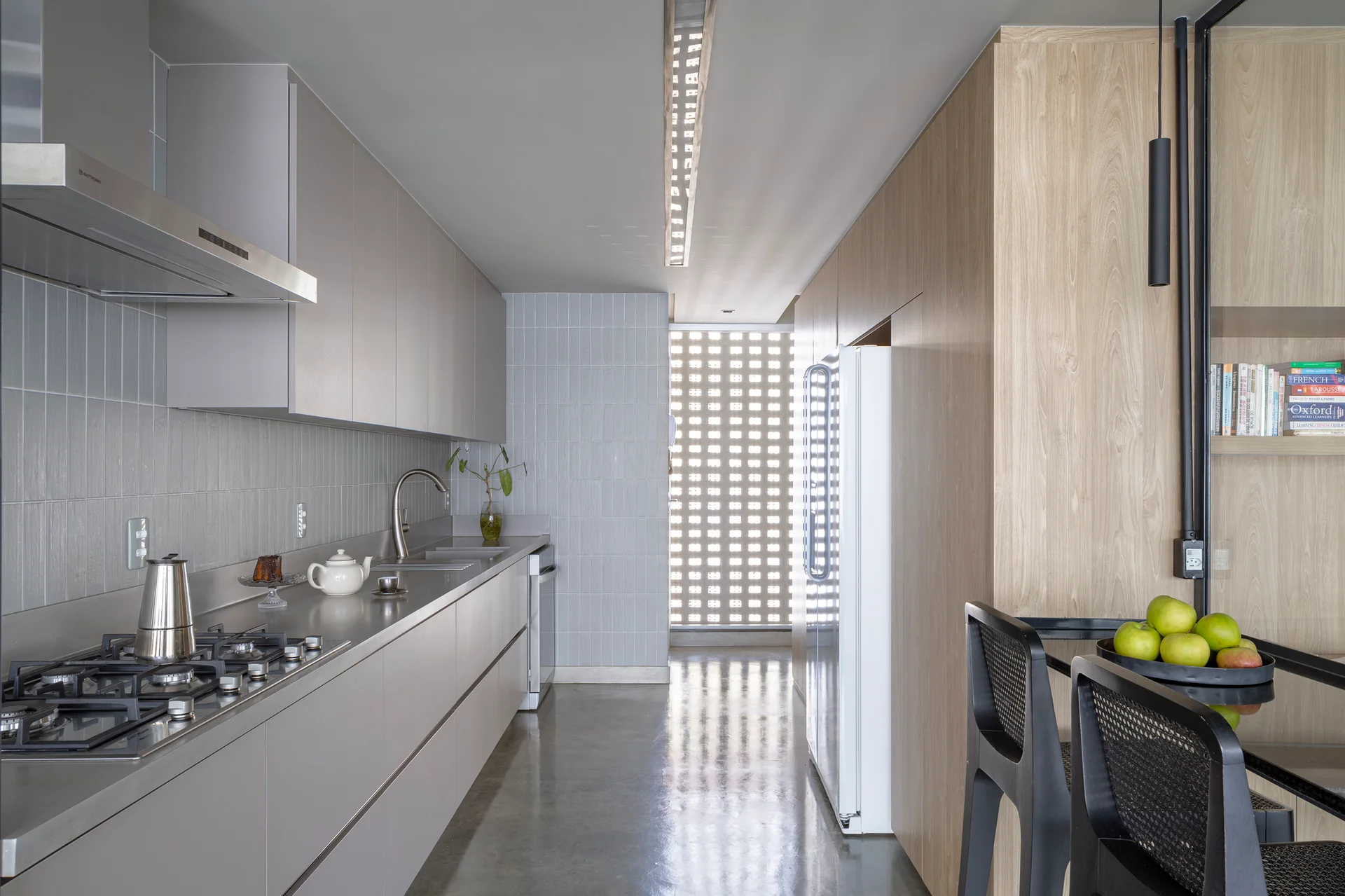 A minimalist galley kitchen with grey cabinets, polished concrete floors, wood wall paneling, and a breeze block window at the far end.