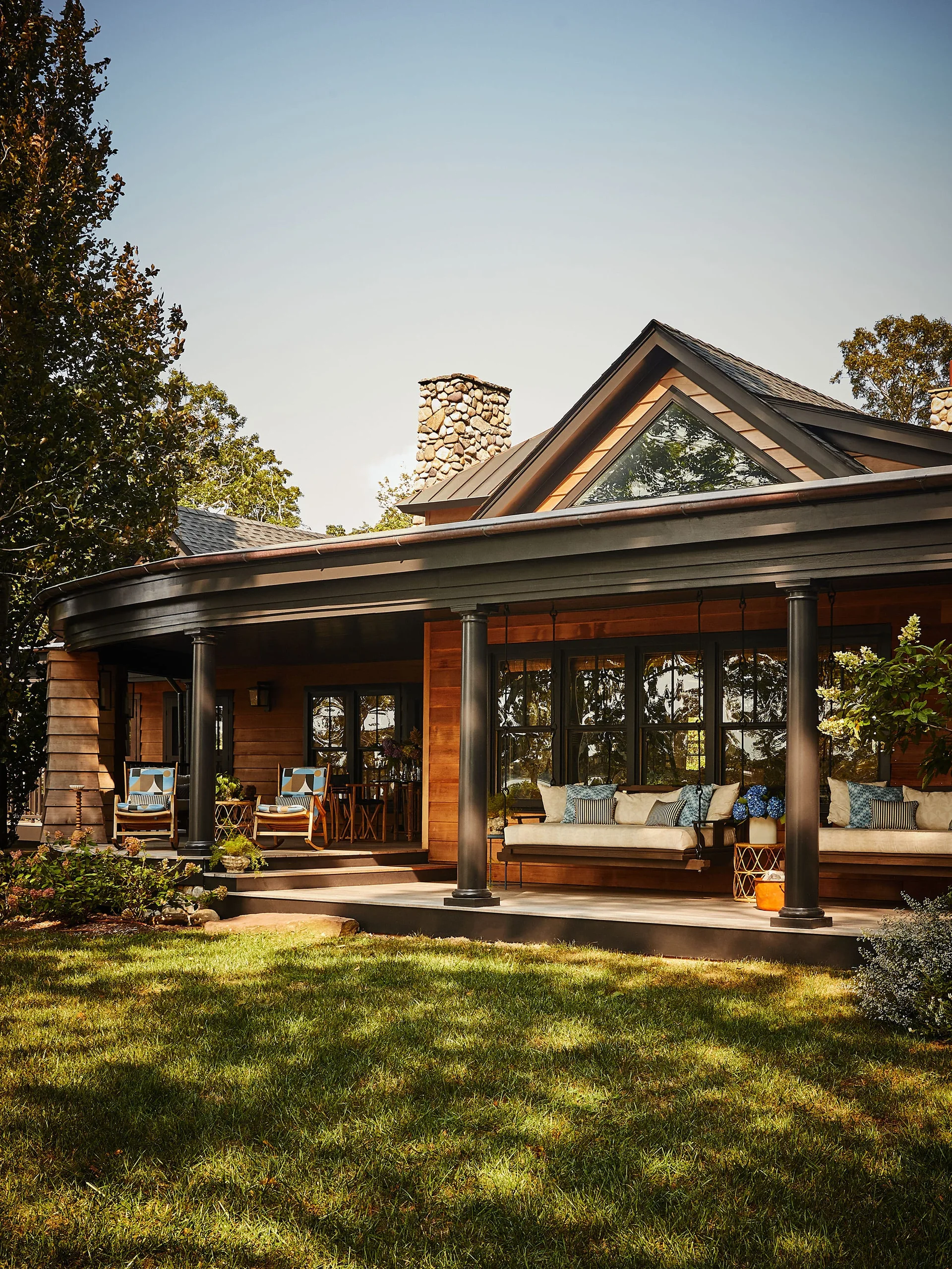 A spacious covered wood porch features two hanging daybeds with blue pillows, dark gray columns, and a stone chimney under a gabled roof.