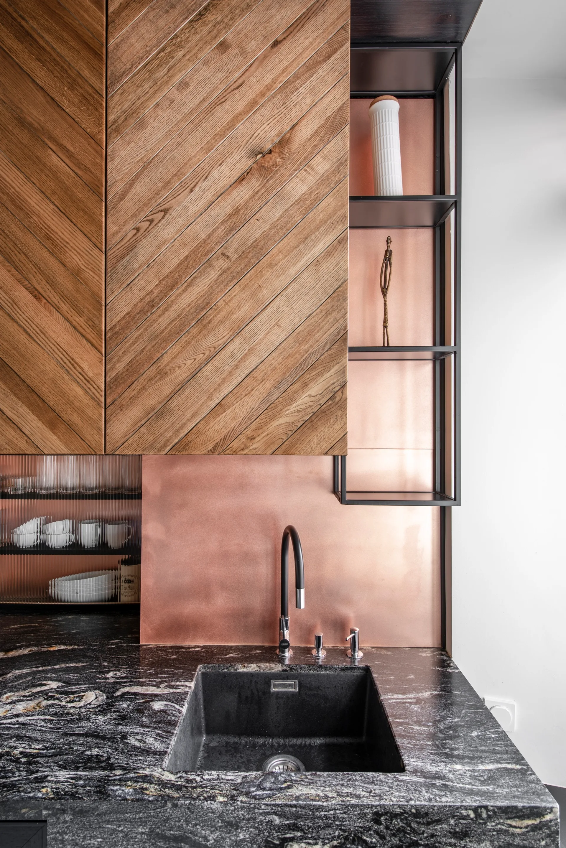 A kitchen sink area features herringbone patterned wood cabinets, a metallic copper backsplash, black stone countertops, and a black faucet.