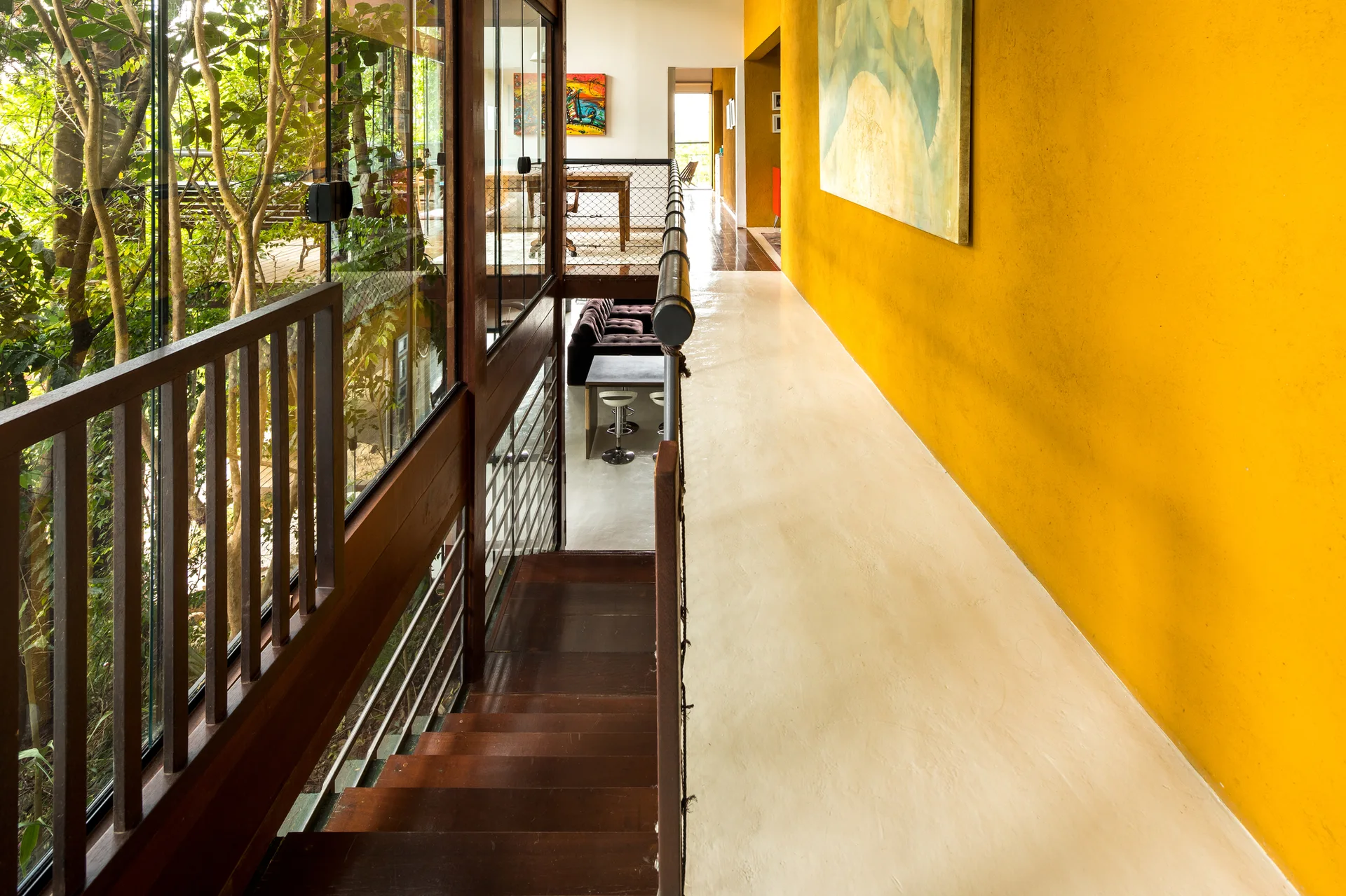 A modern hallway with a bright yellow wall features a polished cream floor, a wooden staircase, and a glass partition showing green trees outside.