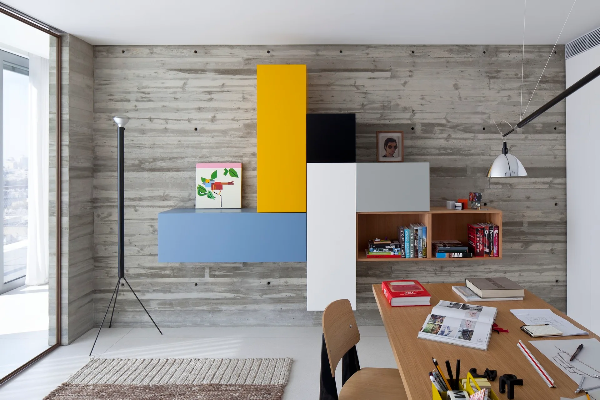 A wooden desk and chair in a home office featuring a textured gray concrete wall with colorful, geometric modular storage boxes mounted on it.
