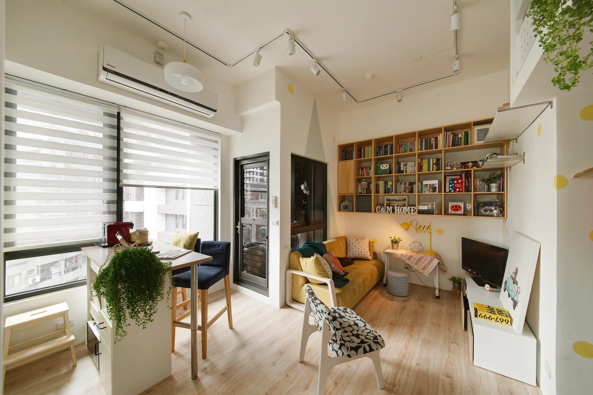 A bright apartment living room features a mustard yellow sofa beneath a large wood bookshelf, a counter-height dining table, and light wood flooring.