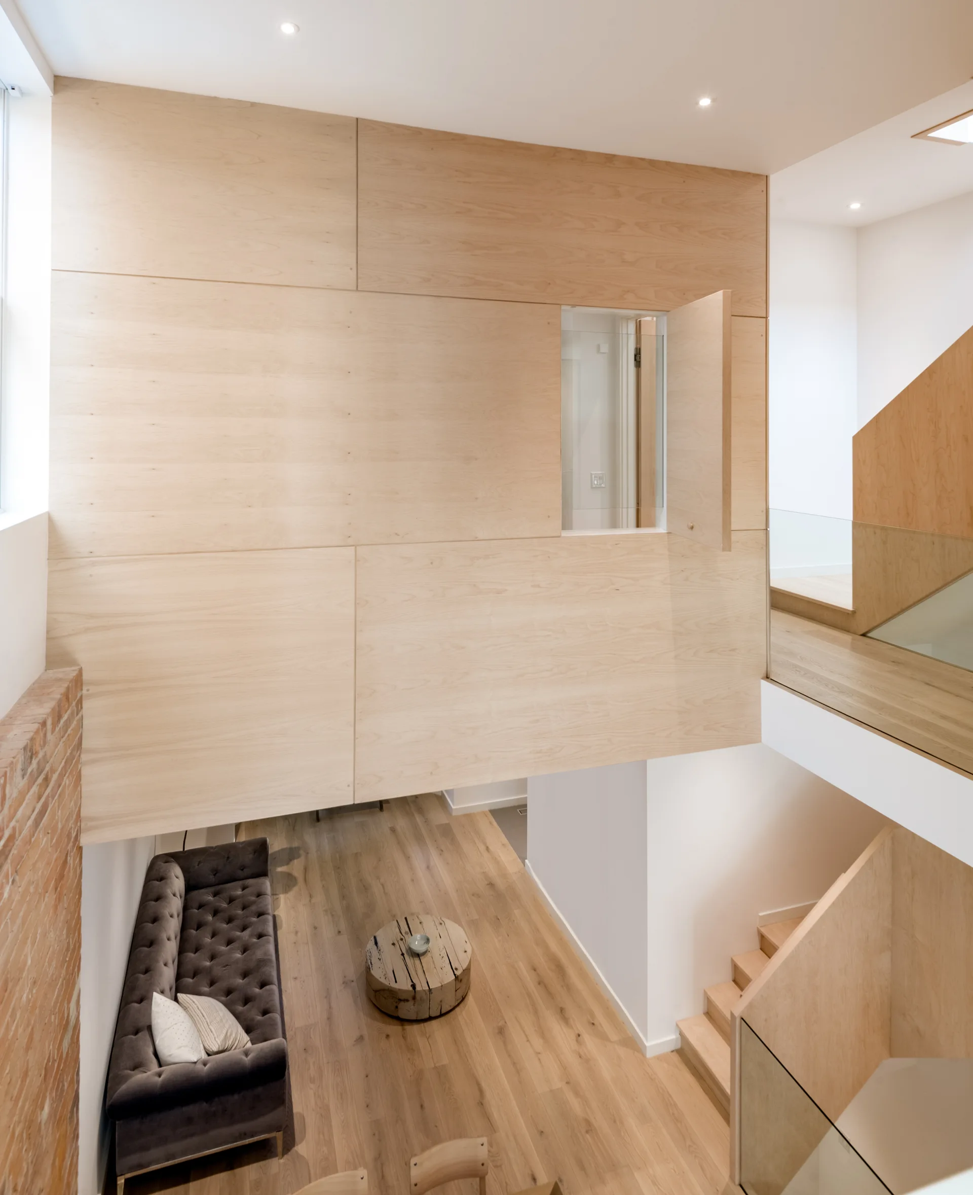 High-angle view of a modern living room featuring a tufted grey sofa, a round wooden coffee table, a glass-railed staircase, and light wood wall panels.
