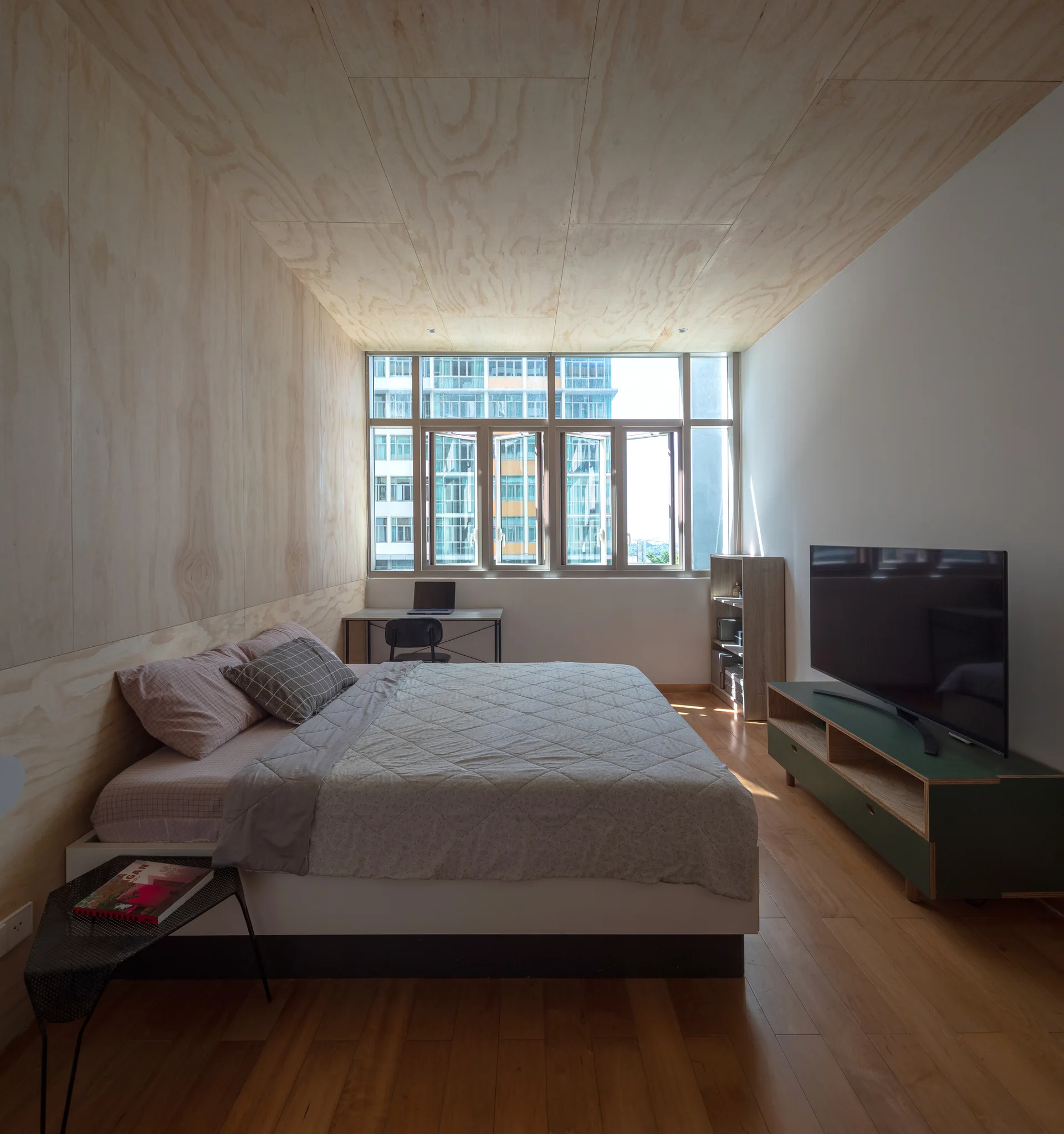 A minimalist bedroom features light plywood wall and ceiling paneling, a large bed with a quilted grey cover, and a forest green television console.
