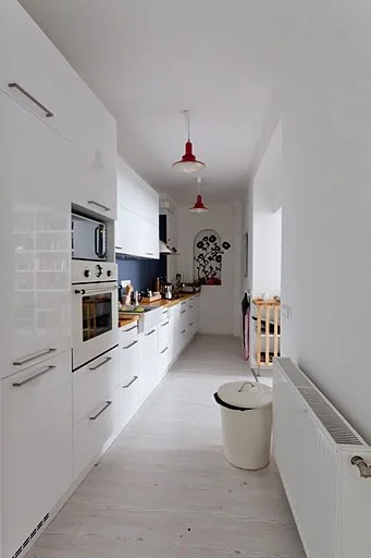 A narrow white galley kitchen with glossy cabinets, wood countertops, built-in appliances, and two hanging red pendant lights.