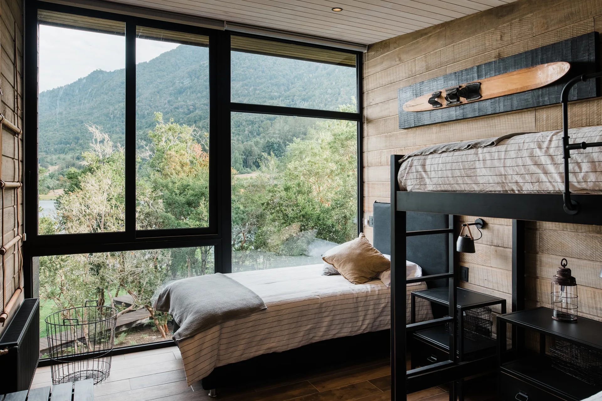 A rustic bedroom with a black metal bunk bed, a low bed by a large window looking out at mountains, and horizontal wood-paneled walls.