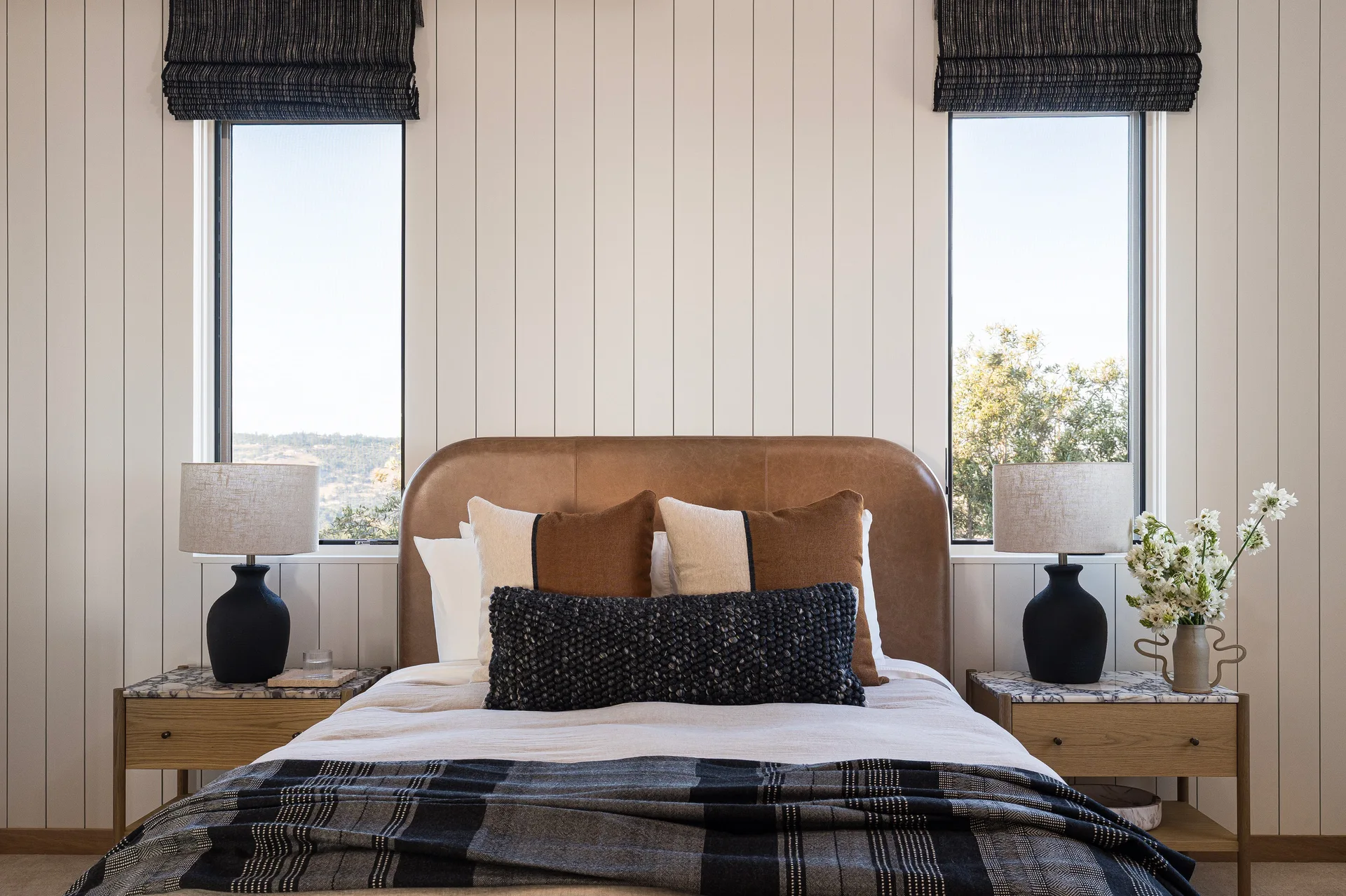 A symmetrical bedroom featuring a tan leather headboard, vertical white paneled walls, two narrow windows, and wood nightstands with black lamps.
