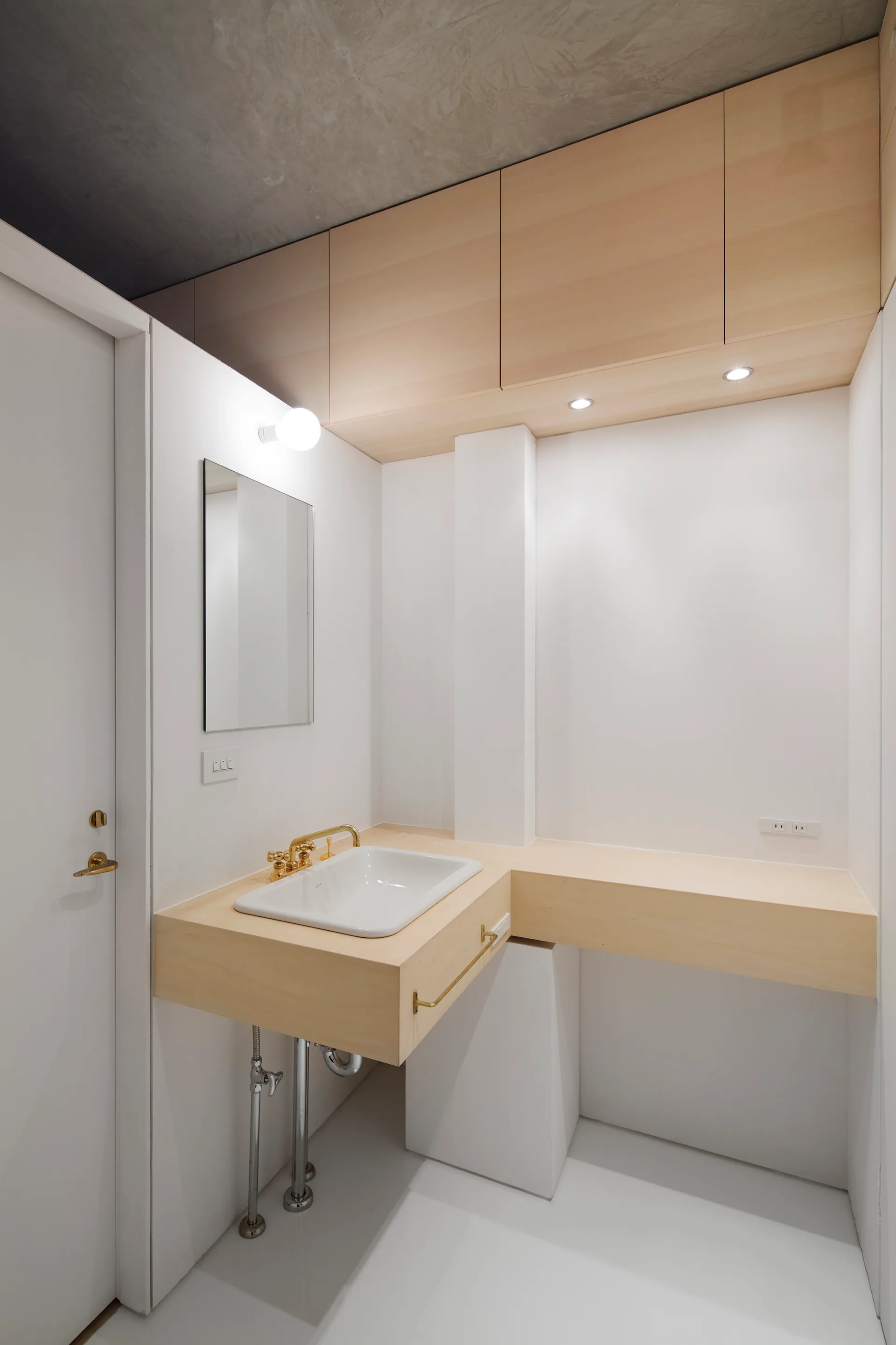 A modern minimalist bathroom featuring a floating light wood vanity, white vessel sink, gold faucet, and a raw concrete ceiling.