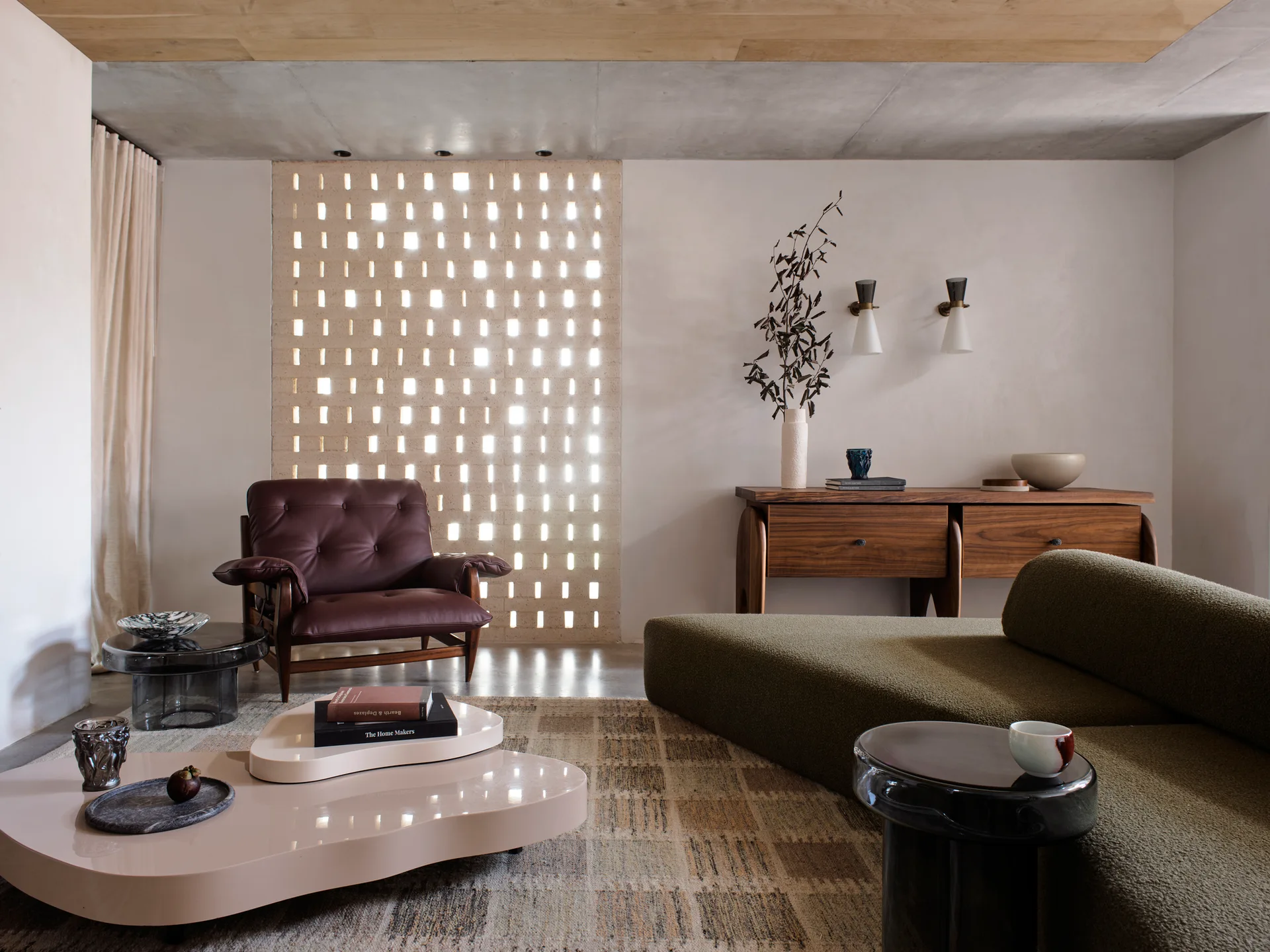 An organic modern living room features an olive green boucle sofa, a tufted brown leather armchair, and a light-filtering perforated brick screen.