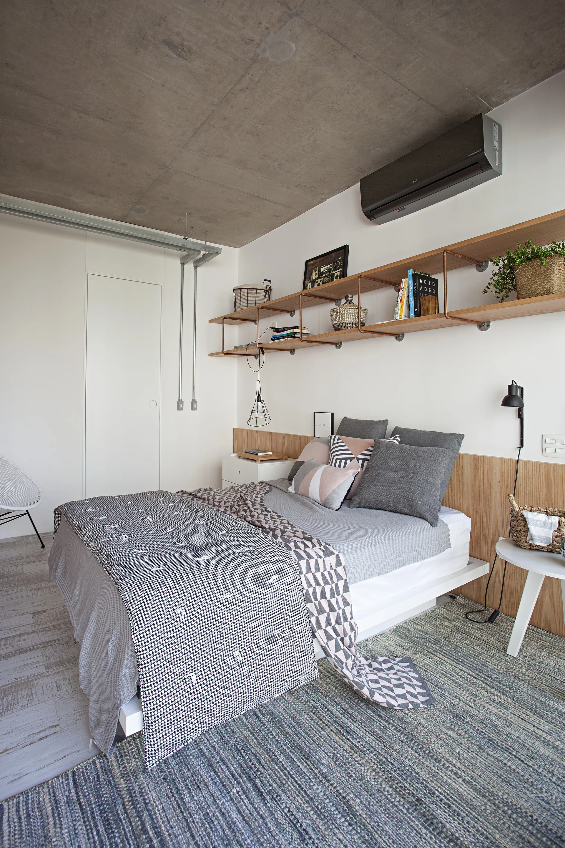 A modern platform bed with grey linens sits against a wooden panel headboard beneath an exposed concrete ceiling with wall-mounted copper pipe shelving.