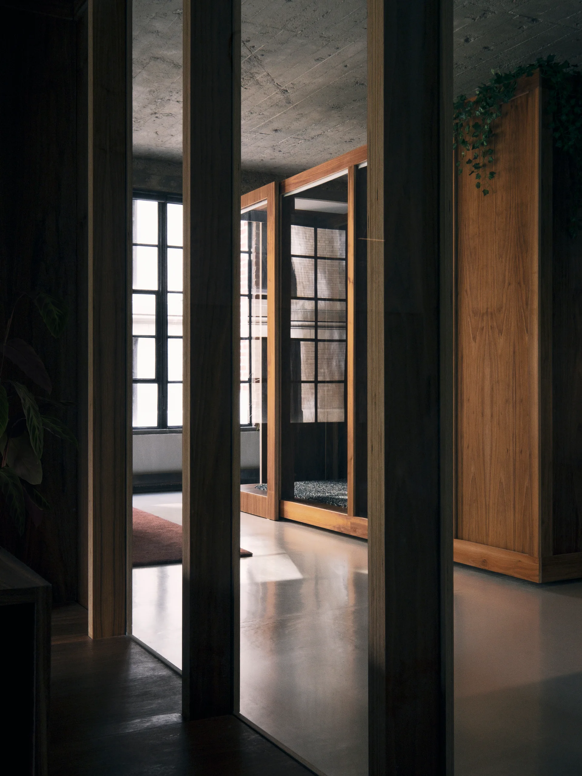 Vertical wooden pillars stand in the foreground of an industrial loft featuring wood-framed glass cabinetry, a concrete ceiling, and grid windows.