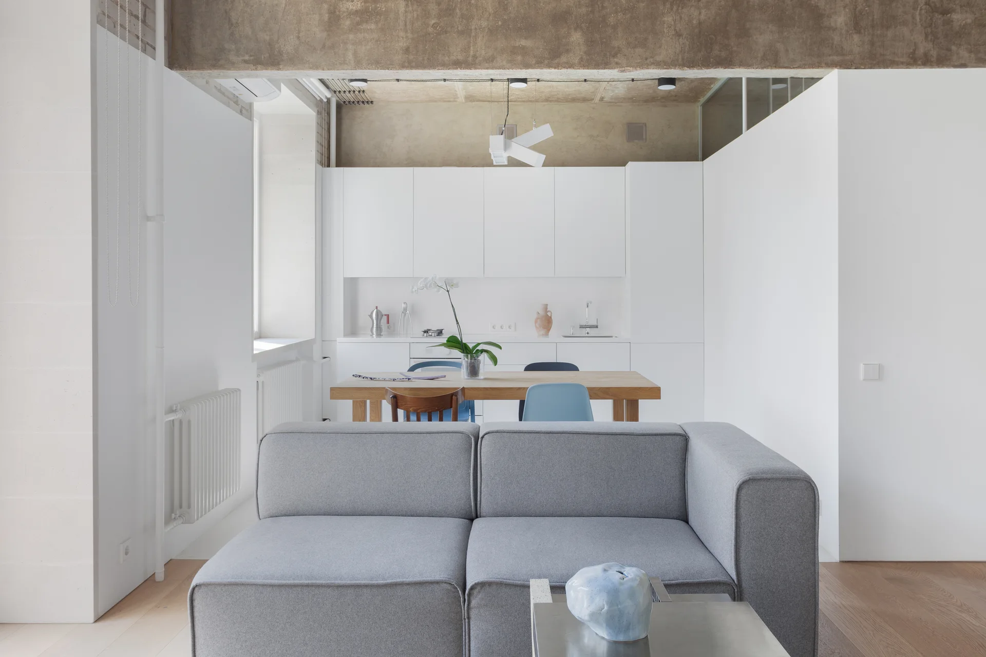 An open-concept living space features a gray sofa, a wood dining table, and minimalist white kitchen cabinets beneath an exposed concrete ceiling beam.