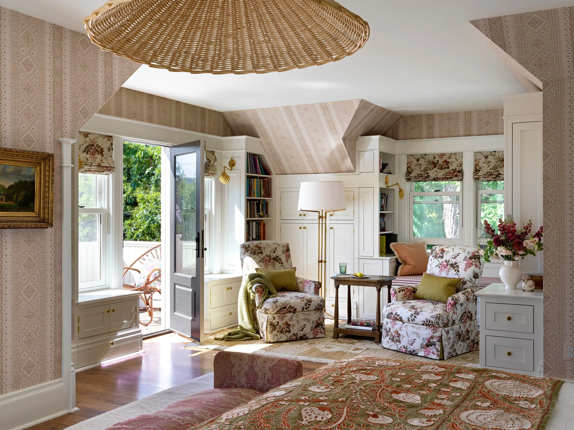 A cozy cottage bedroom features floral patterned armchairs, custom white built-in shelving, and geometric wallpaper under a sloped ceiling.