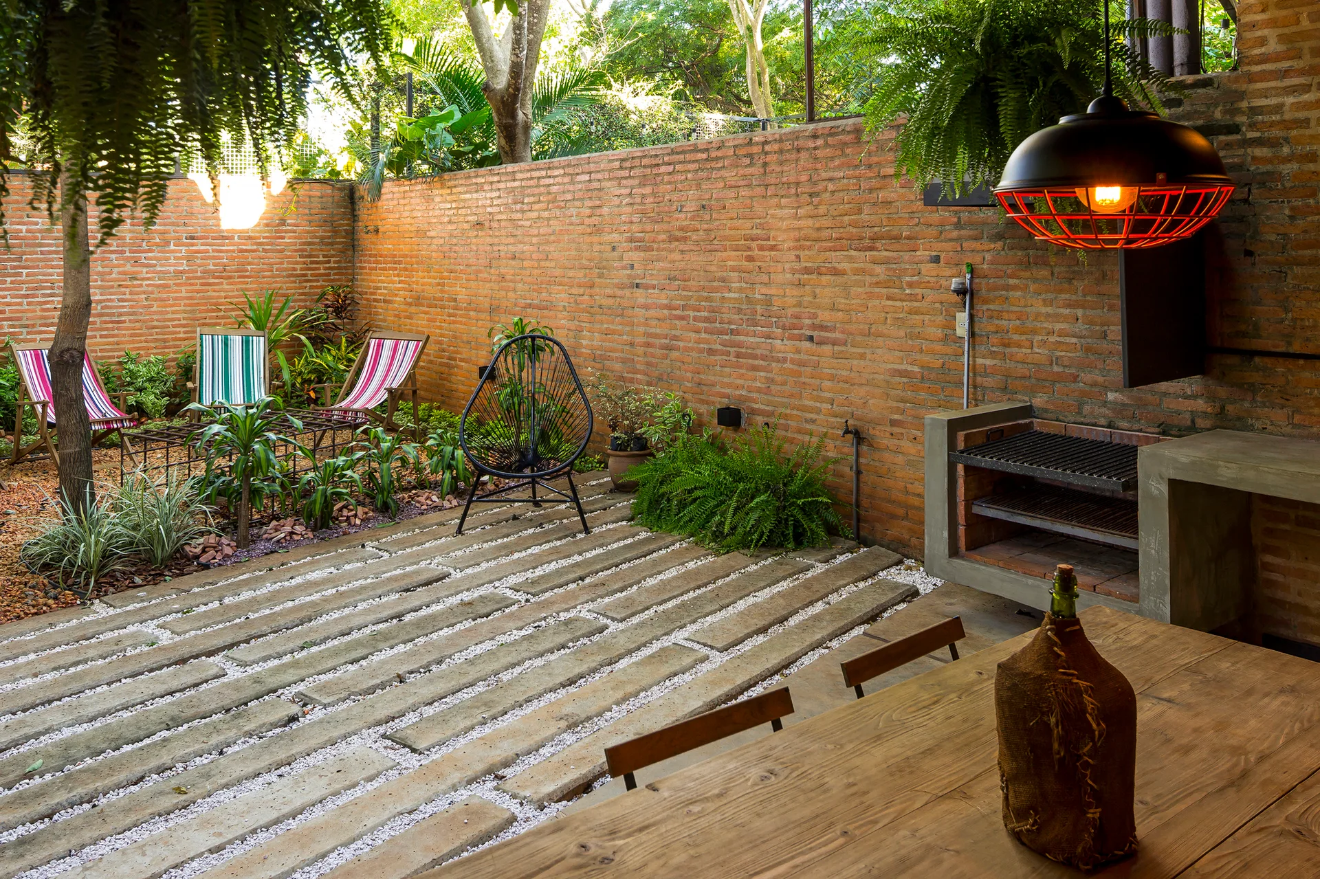 An outdoor backyard patio features an exposed brick wall, concrete plank pavers set in white gravel, a built-in concrete grill, and a wooden dining table.