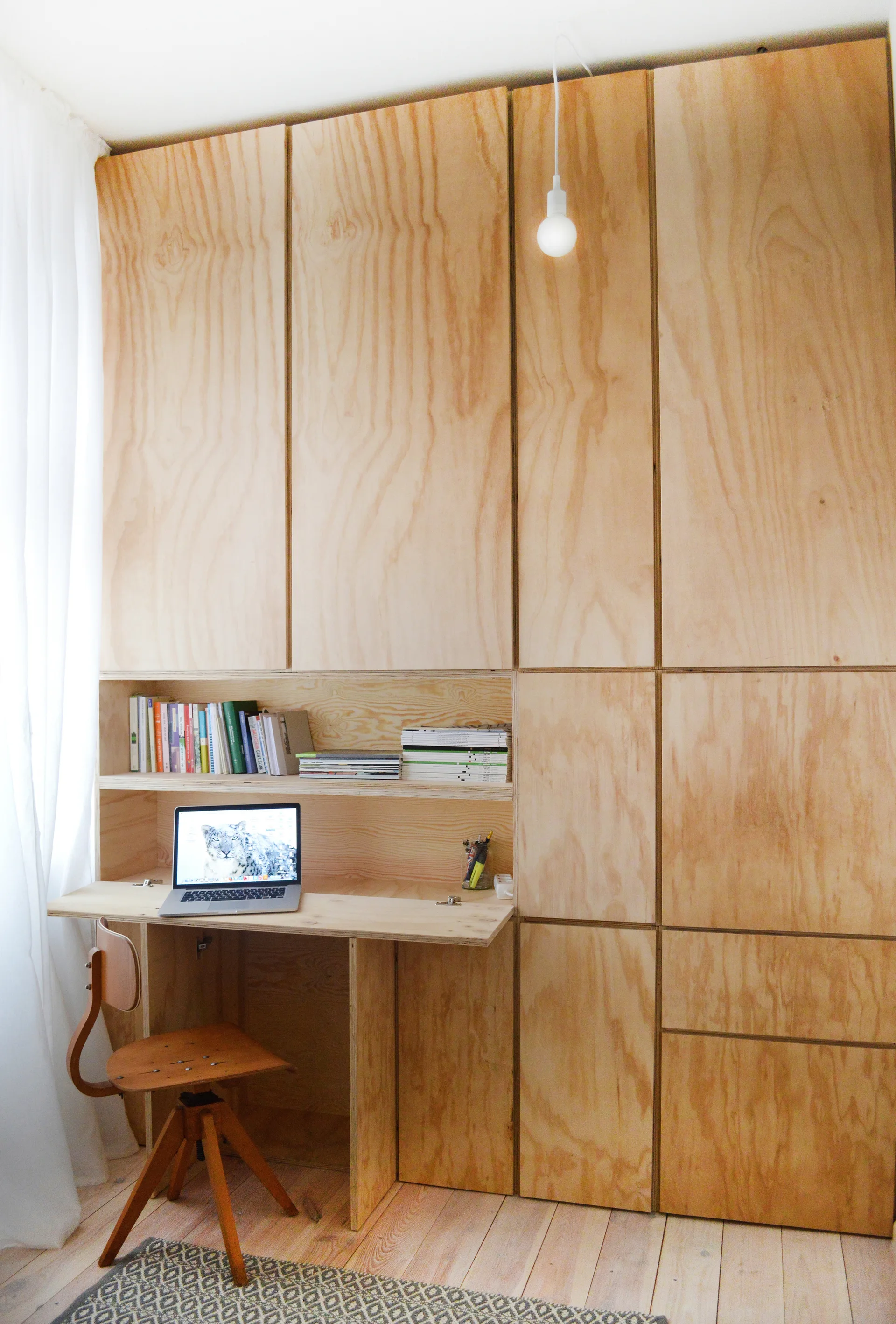 A home office workspace features a custom plywood desk integrated into floor-to-ceiling cabinetry beside a wooden swivel chair and sheer white curtains.