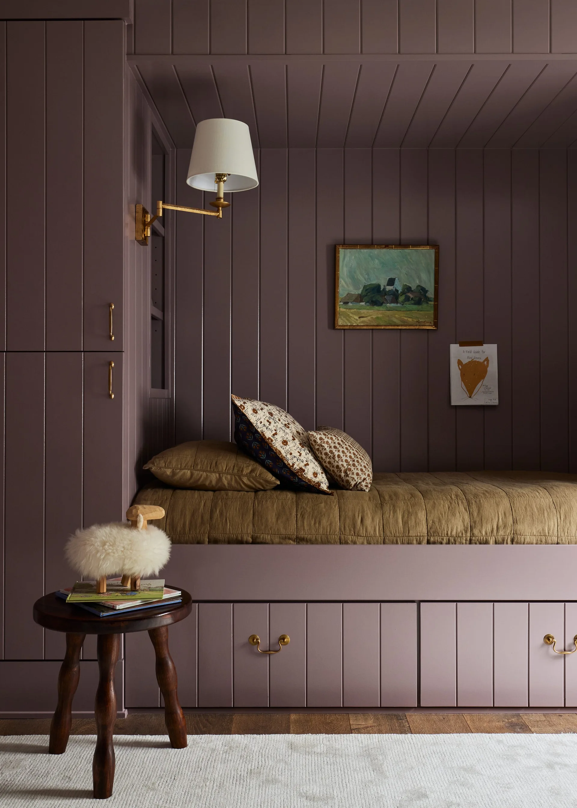 A built-in daybed nook painted mauve features vertical wood paneling, integrated storage drawers, a brass wall sconce, and earthy-toned bedding.