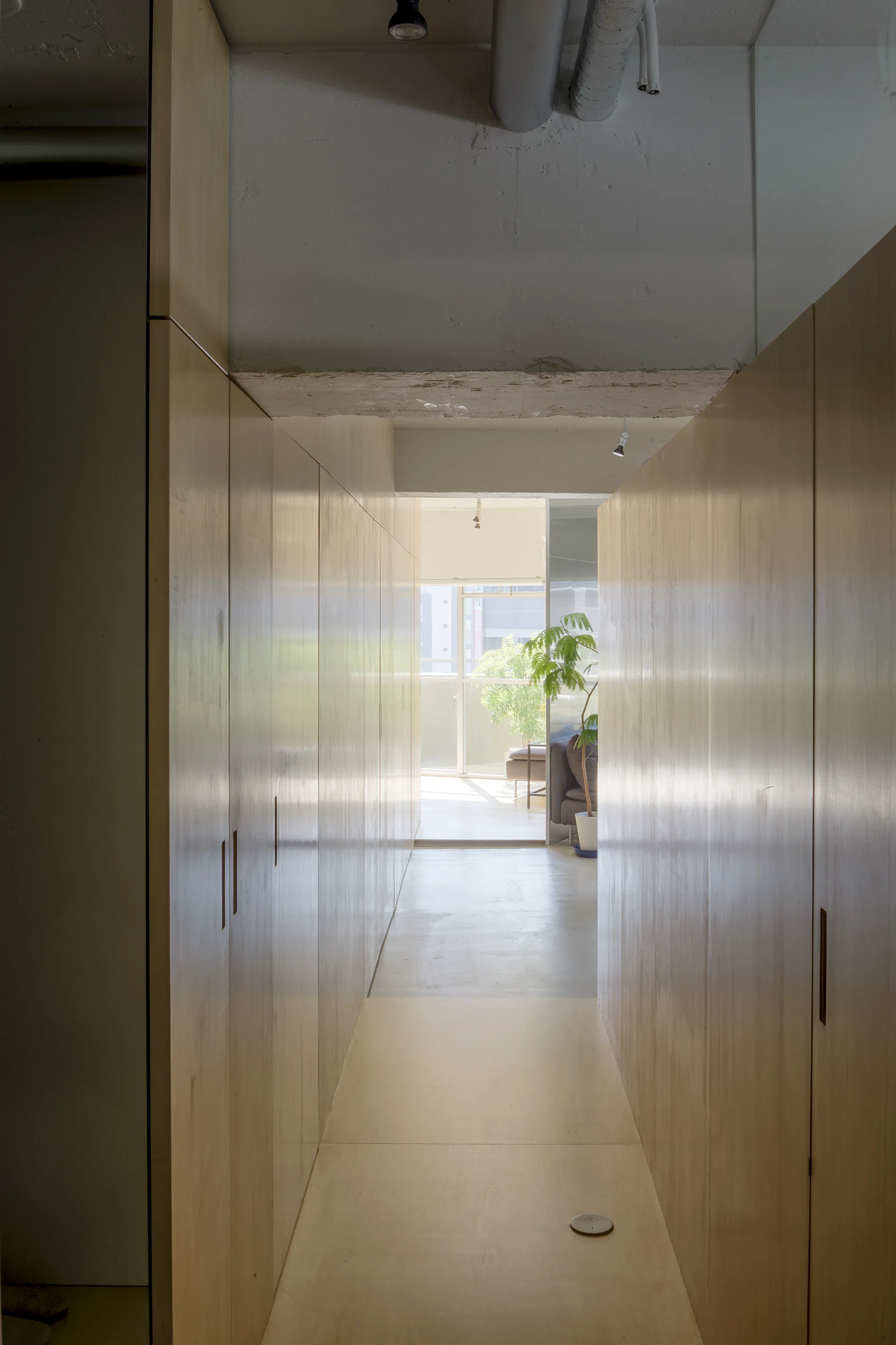 A narrow interior hallway lined with floor-to-ceiling light wood cabinetry, featuring a smooth grey floor and an exposed concrete ceiling.