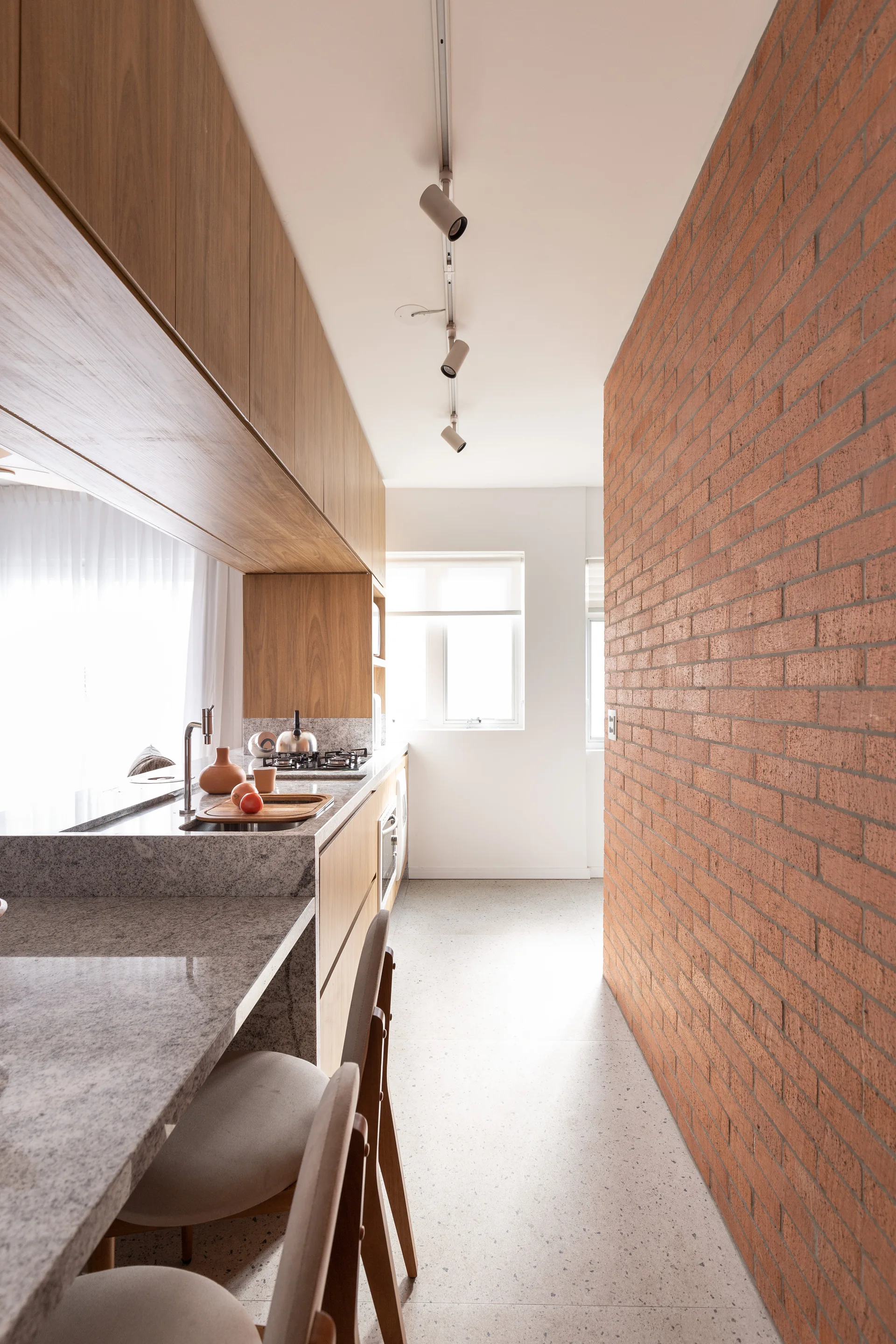 A galley kitchen features natural wood cabinets, a grey stone countertop with bar stools, a red brick accent wall, and ceiling track lighting.
