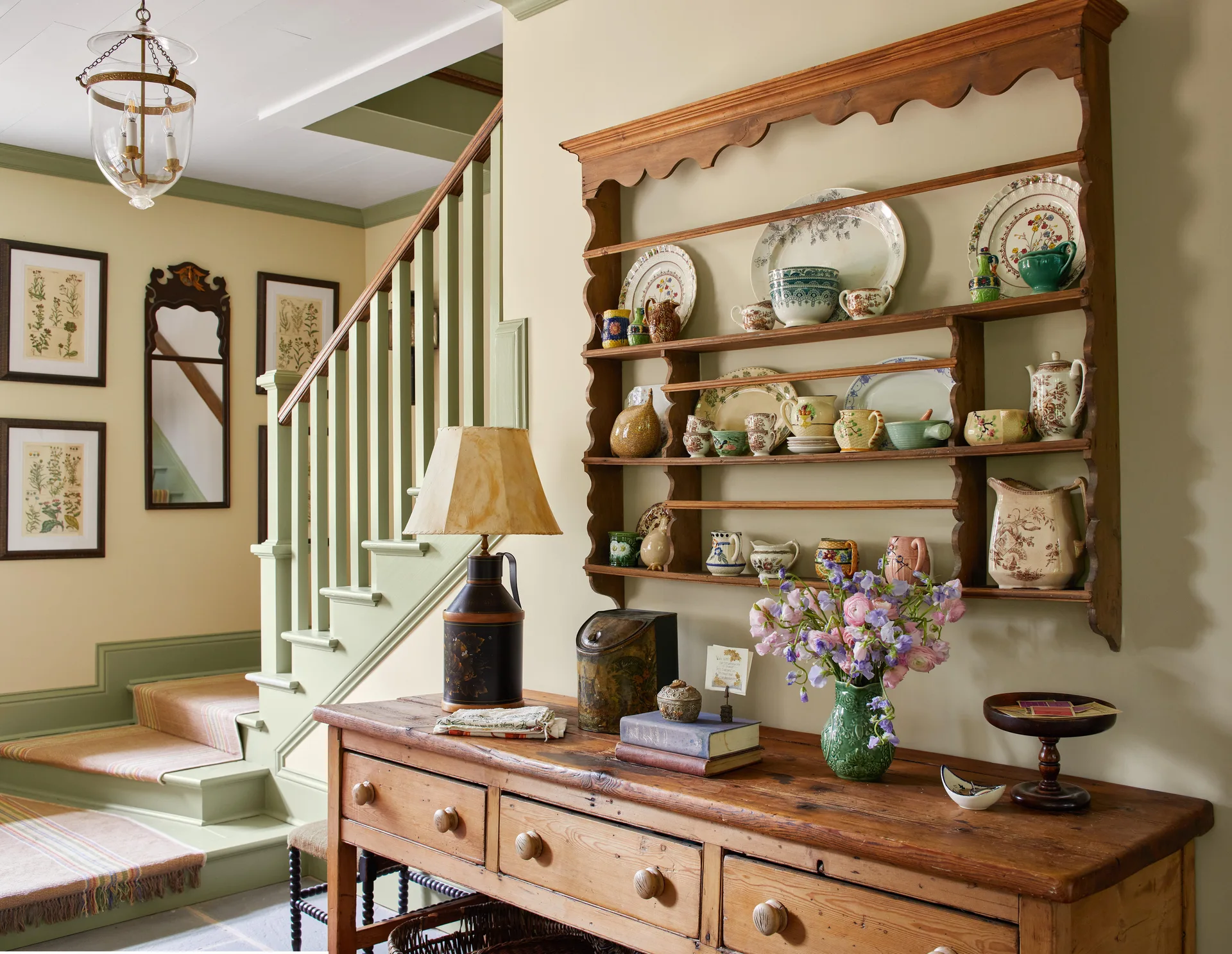 A cottage entryway features a wooden console table beneath a wall-mounted plate rack, next to a staircase with sage green painted wood balusters.