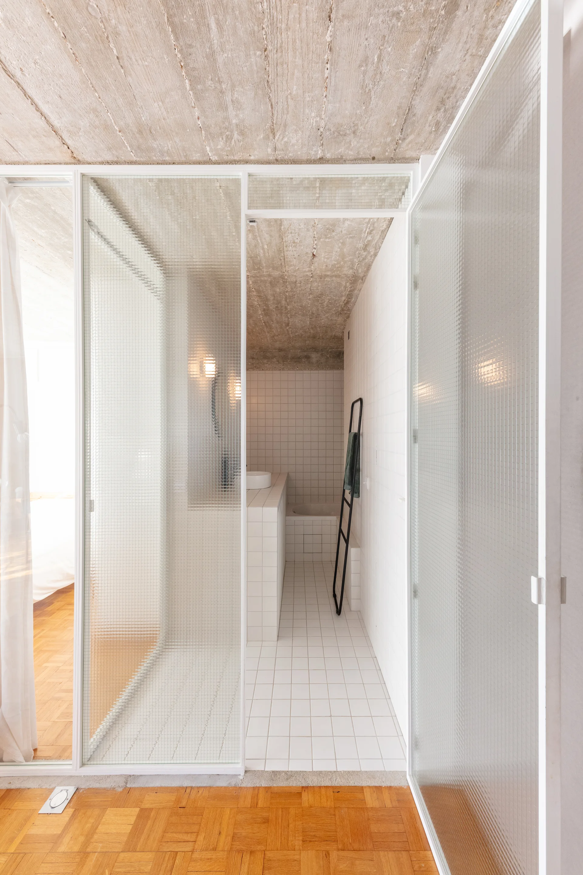 A minimalist bathroom features white square tiles on the walls and floor, a textured glass partition wall, and an exposed concrete ceiling.