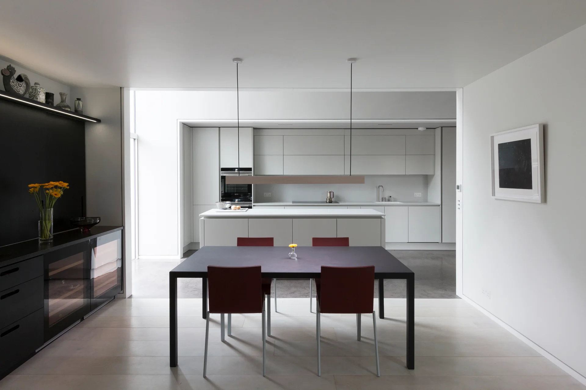 A minimalist open-plan dining area features a black table with maroon chairs, leading to a modern white kitchen with handleless cabinets.