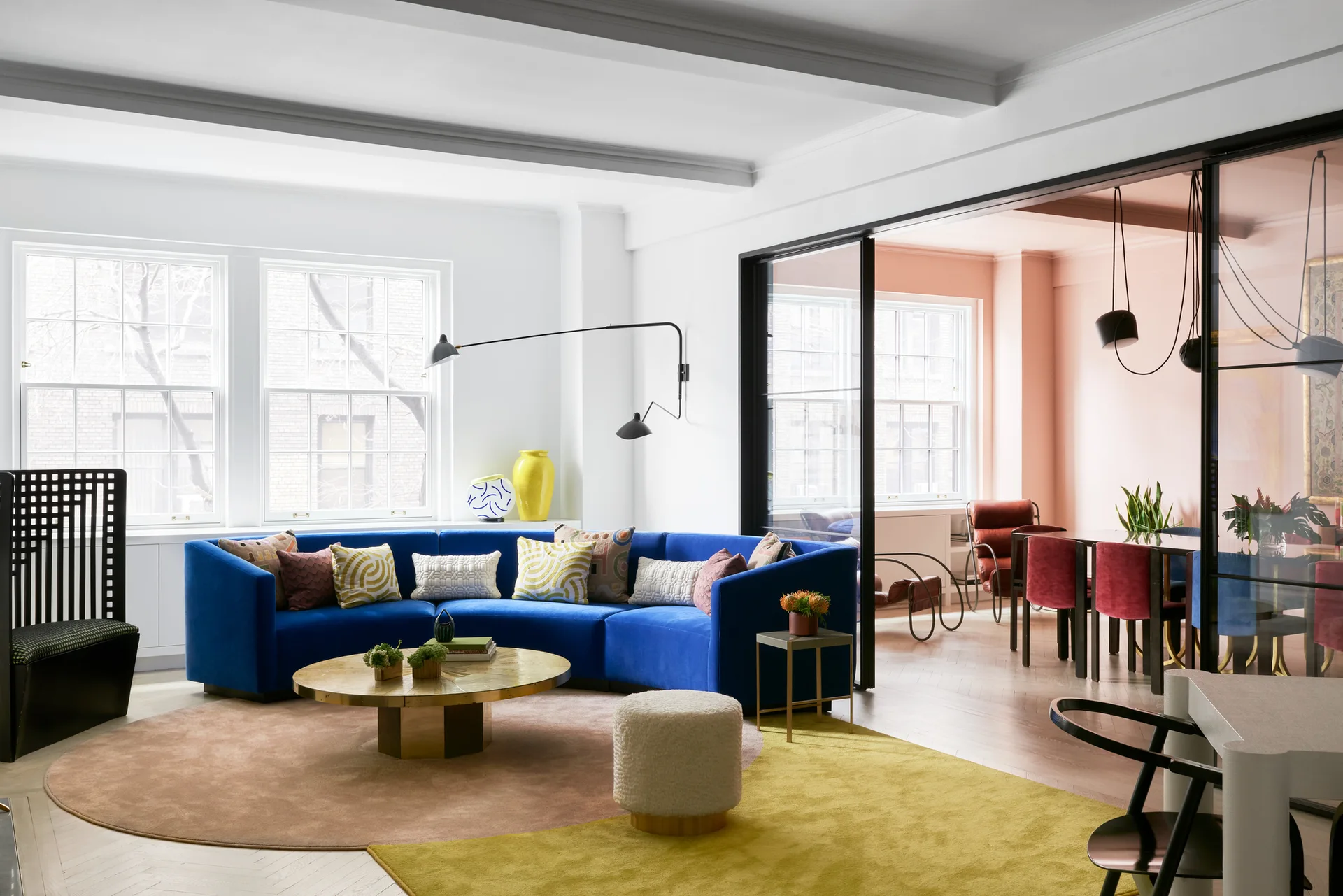 A living room with a curved blue velvet sofa and a gold coffee table next to a black-framed glass partition looking into a pink dining area.