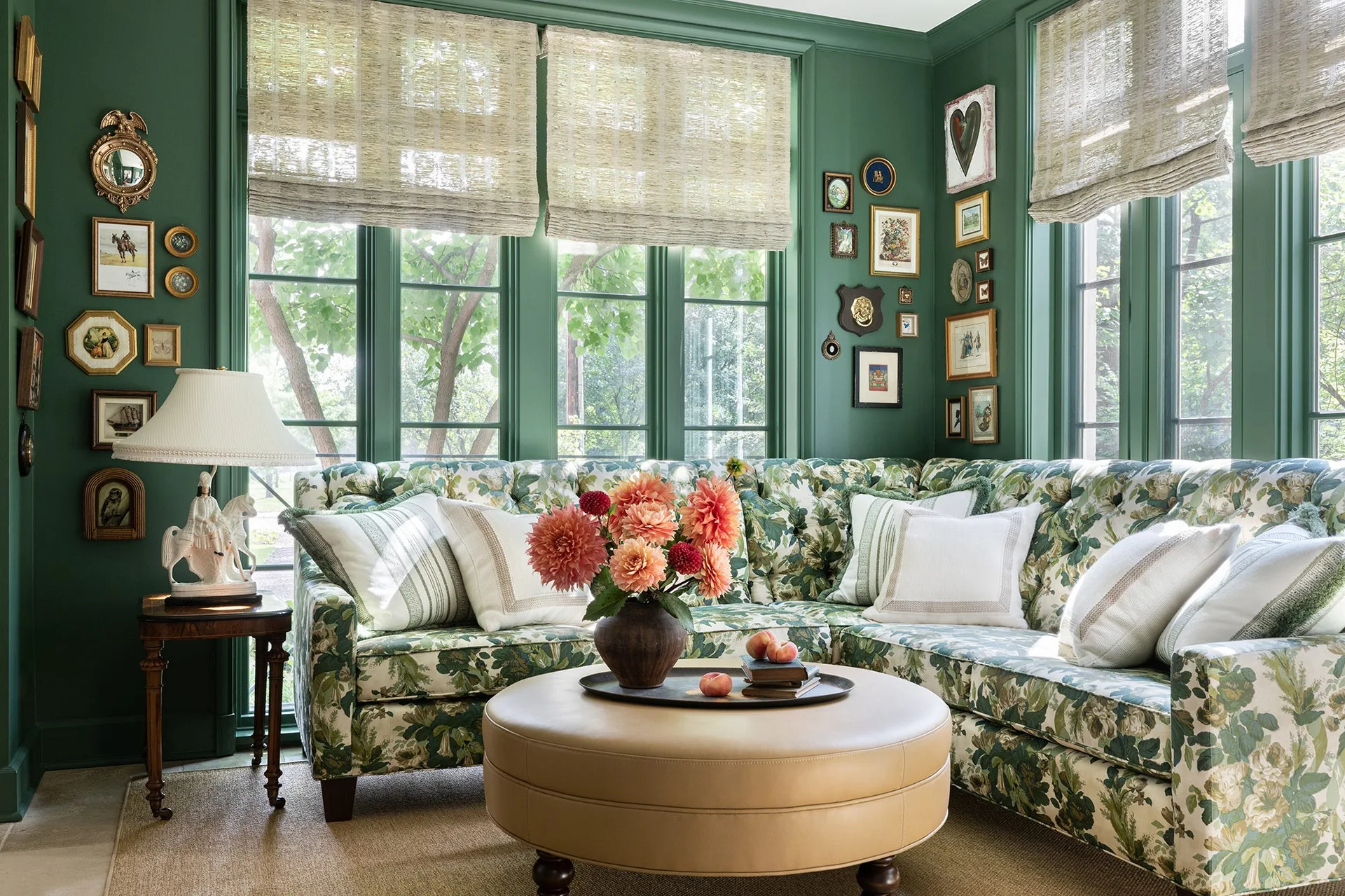 A green living room features a large floral print sectional sofa, a round tan leather ottoman, and woven roman shades covering tall windows.