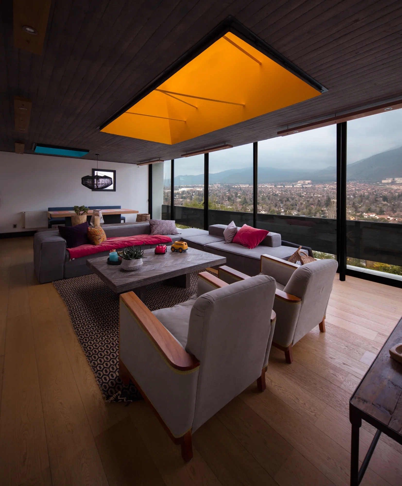 A modern living room features a grey sectional sofa, two armchairs, a patterned rug, and a distinctive bright yellow skylight in a dark wood ceiling.