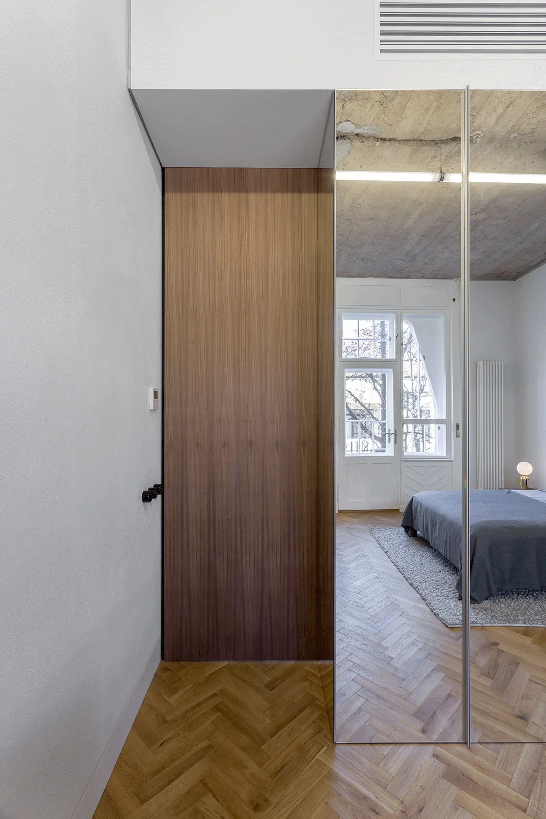 A modern bedroom with herringbone wood floors, a warm wood flat-panel door, and a mirrored wardrobe reflecting the bed.