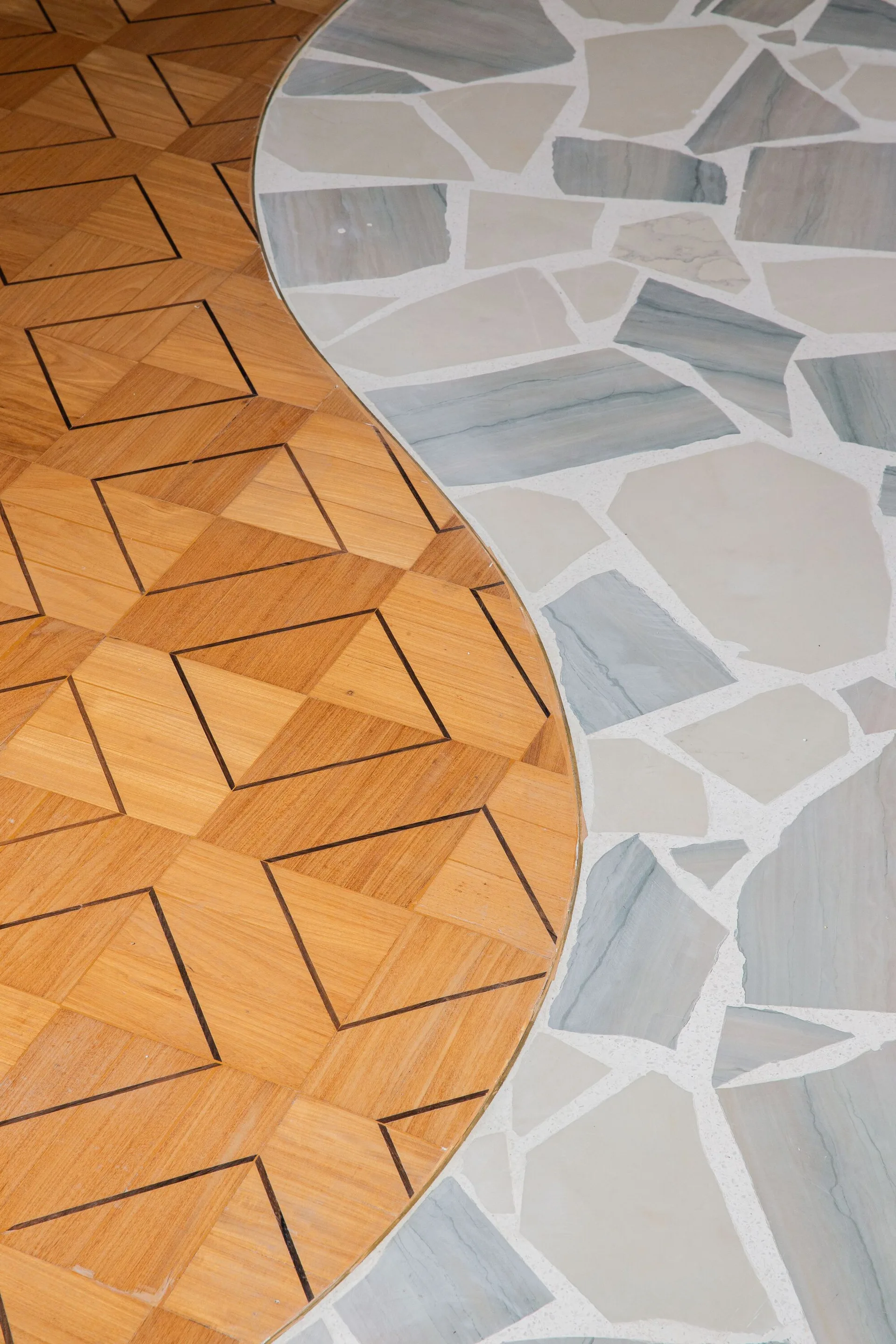 A curved metal inlay separates warm geometric wood parquet flooring from cool gray and white irregular stone mosaic tile.