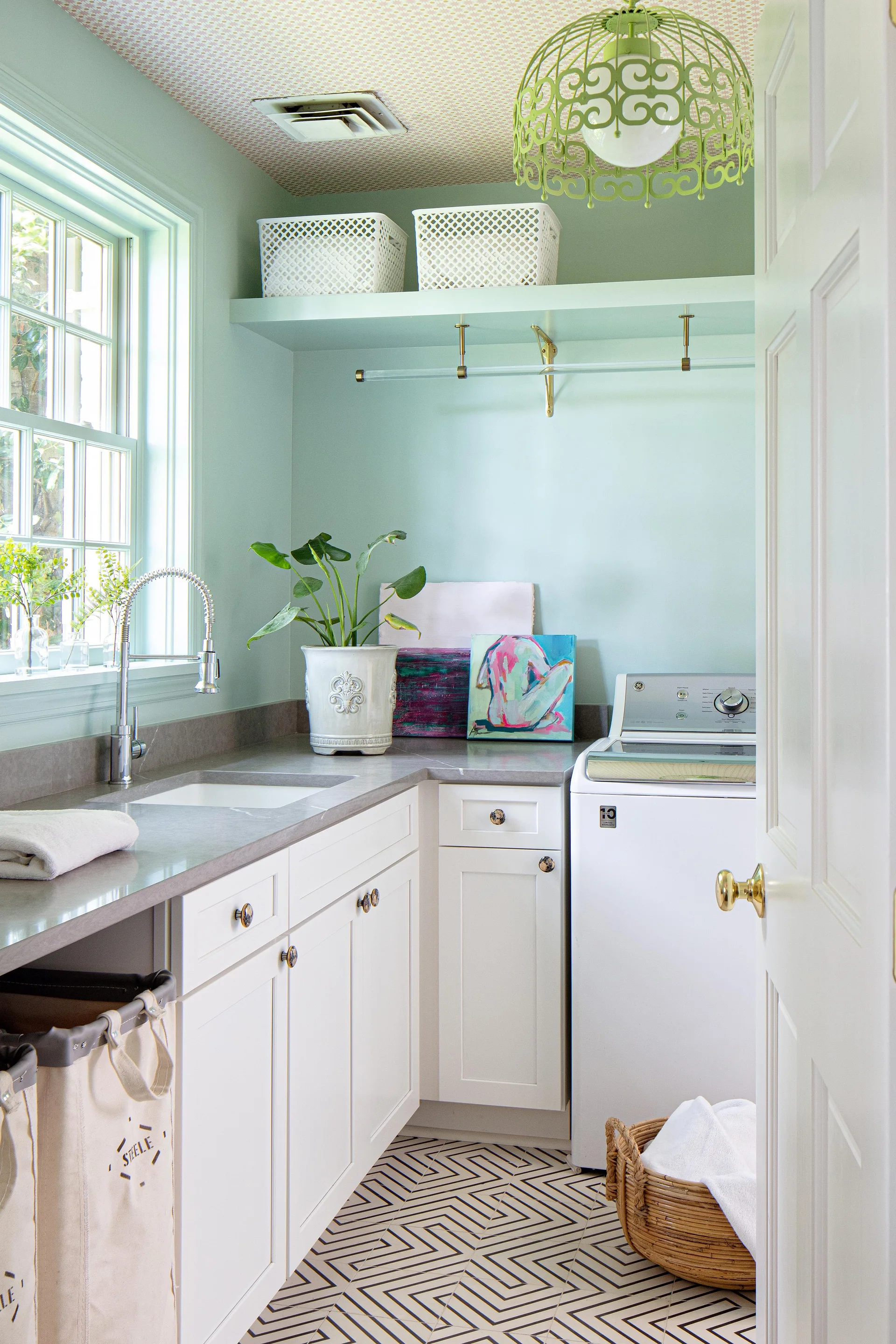 A bright laundry room with mint green walls, white cabinetry, a patterned ceiling, and black and white geometric tile flooring.